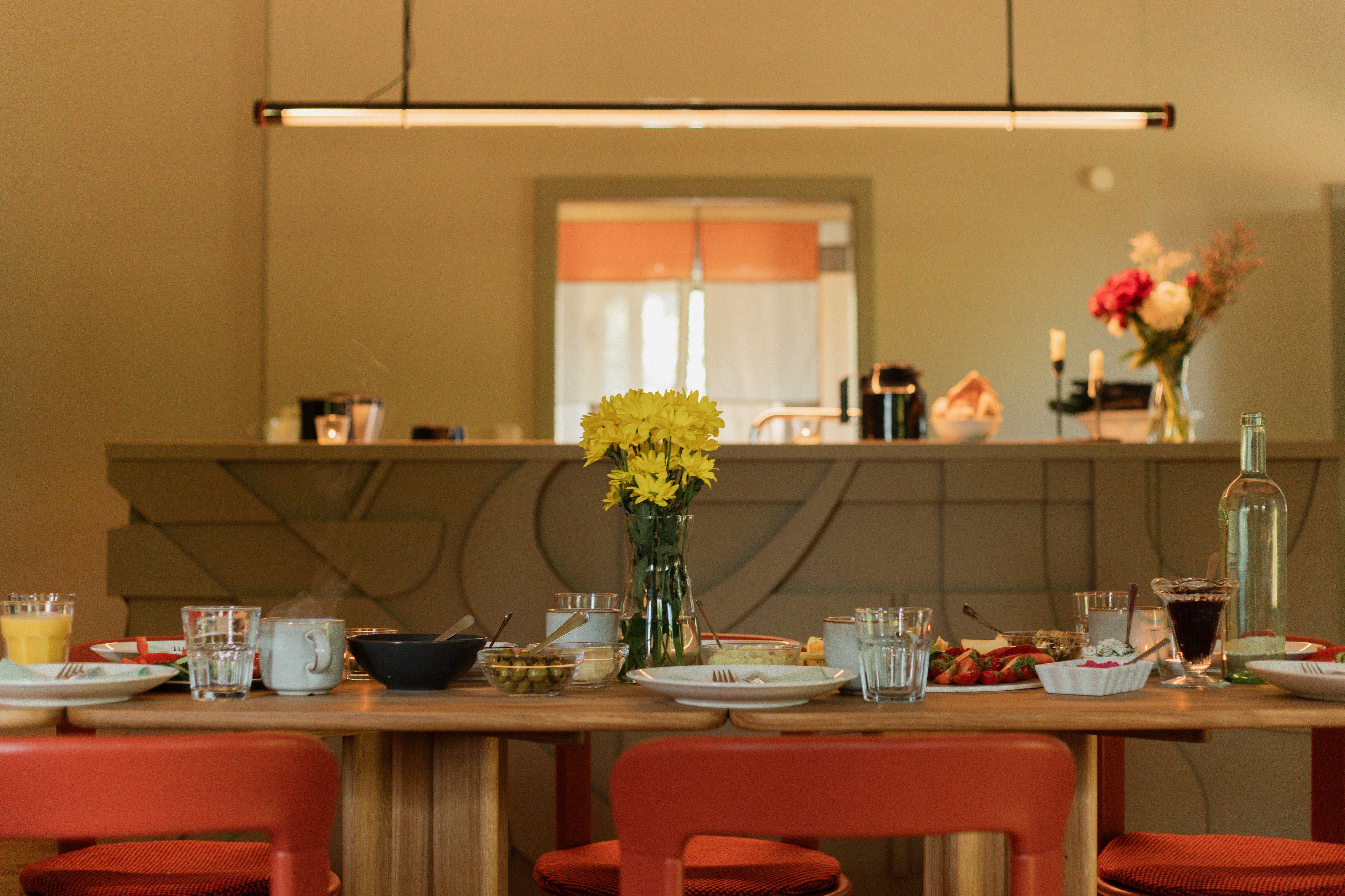 A dining table set for a meal with red chairs, yellow flowers, and various dishes. A modern kitchen counter and soft lighting are in the background.