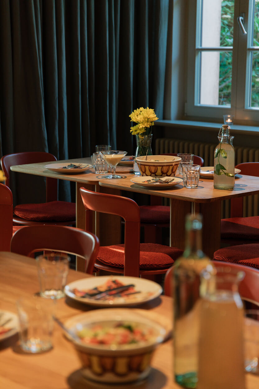 A dining table set with dishes, glasses, and a bottle, featuring a vase of yellow flowers. Red chairs surround the table near a window.