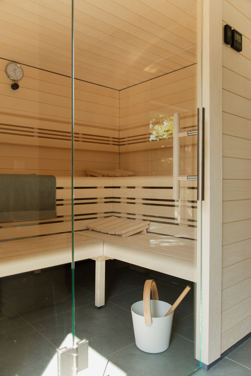 A modern wooden sauna with a glass door, featuring a bench, a towel, a clock, and a bucket with a ladle on the floor.