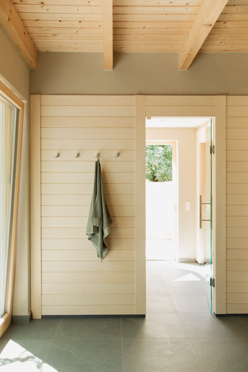 Minimalist room with light wood paneling, a towel hanging on hooks, and an open door leading outside. Sunlight streams through a large window.
