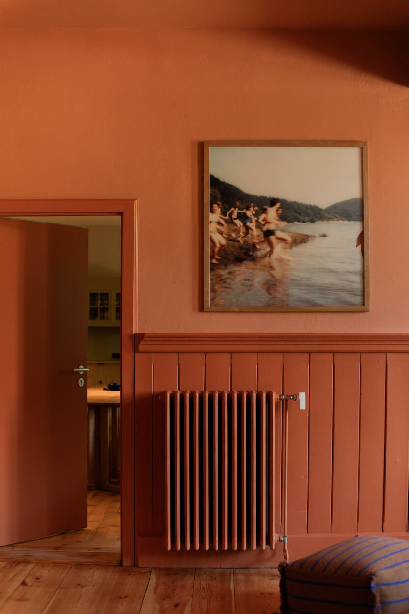 Room with terracotta walls, a radiator, and a large framed photo of people by a lake. An open door reveals a glimpse of a kitchen.