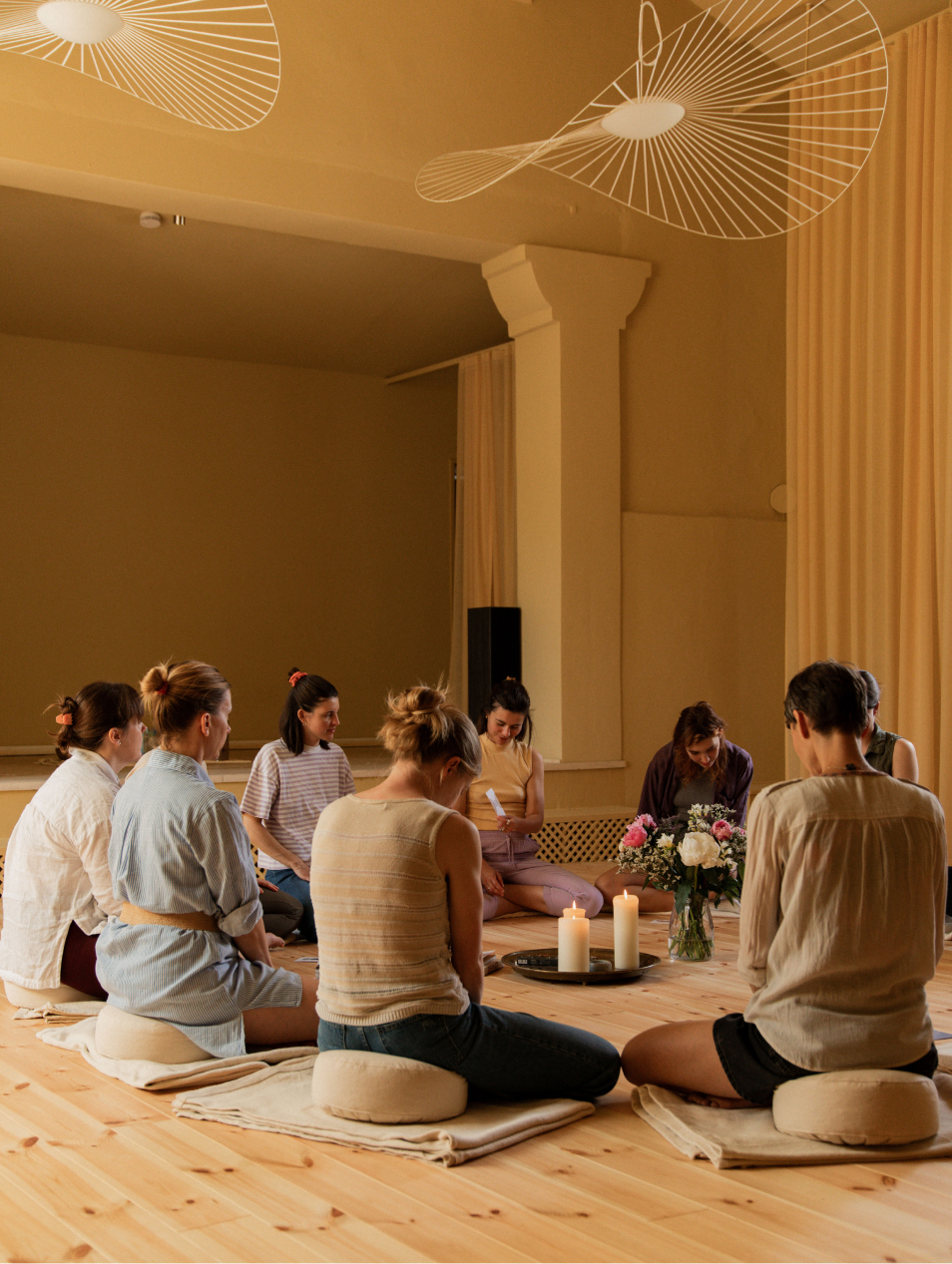 A group of people sitting in a circle on cushions in a serene room, with candles and flowers in the center.