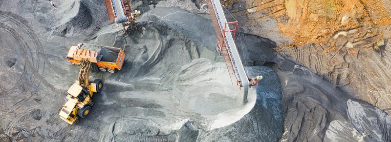 “Aerial view of a gold mining site with heavy machinery, including an excavator loading rocks into a truck and a conveyor system, illustrating the process of gold extraction.