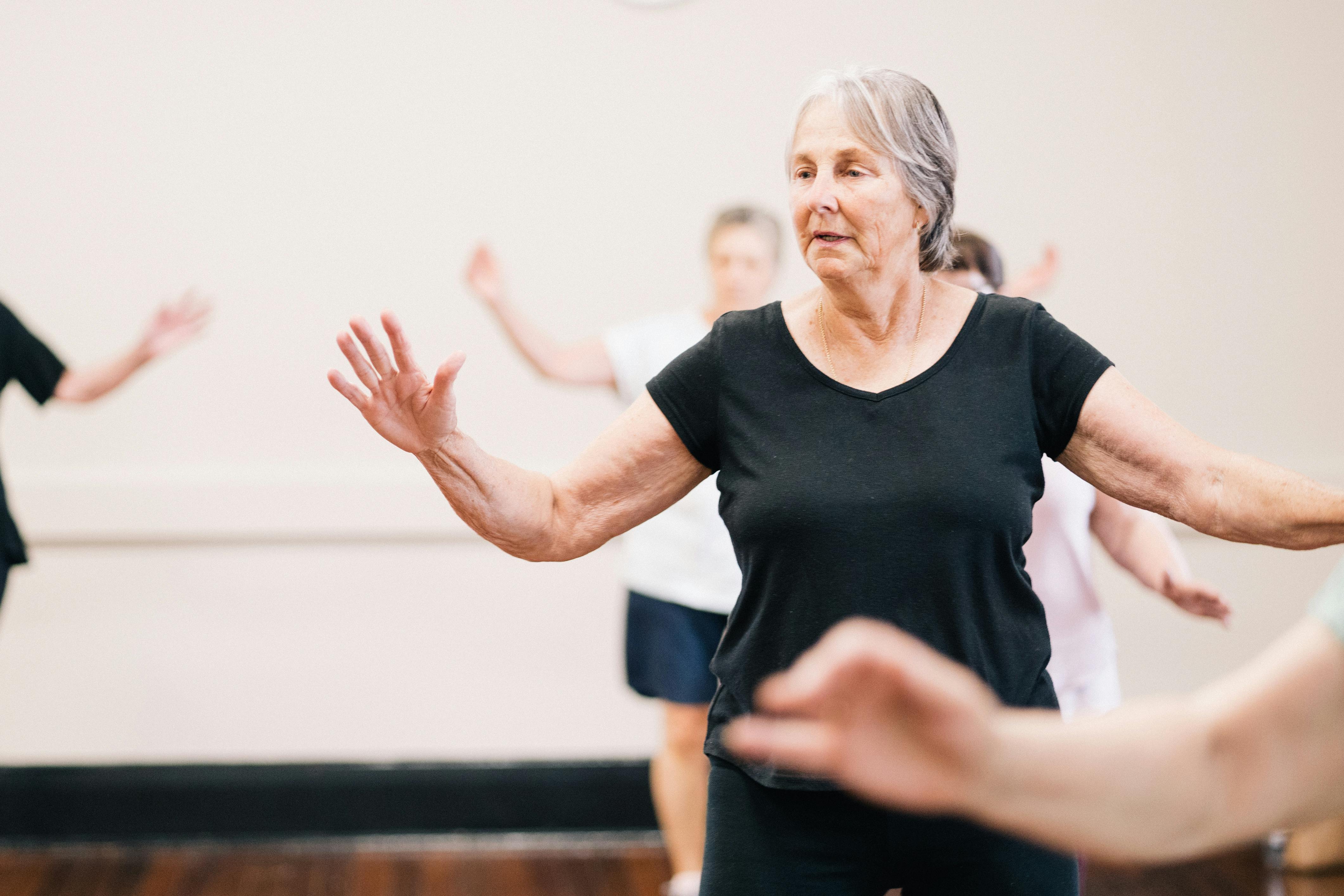 Keeping physically active is important for maintaining mobility, independence, confidence, and in reducing falls risk Senior woman in a black shirt with arms outstretched taking part in a group exercise class