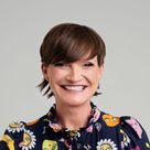 Smiling person with short brown hair wearing a dark blouse with colorful cartoon faces and floral designs, against a plain background.