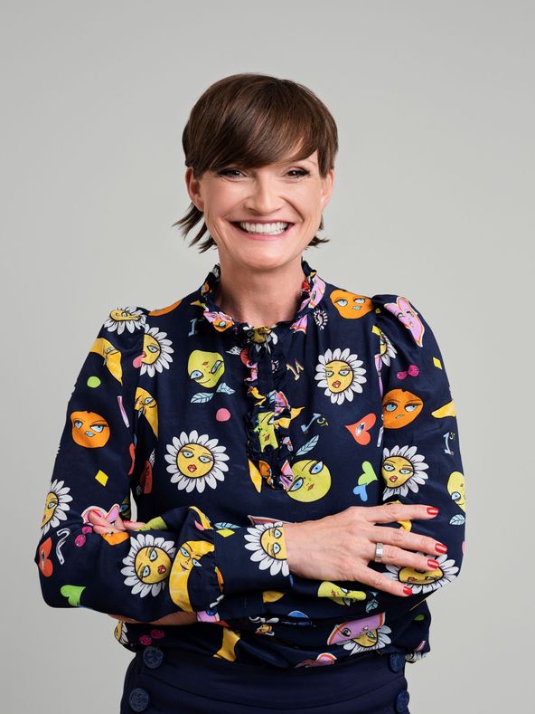 Smiling person with short hair wearing a colorful floral blouse and dark pants against a plain gray background. Arms are crossed.