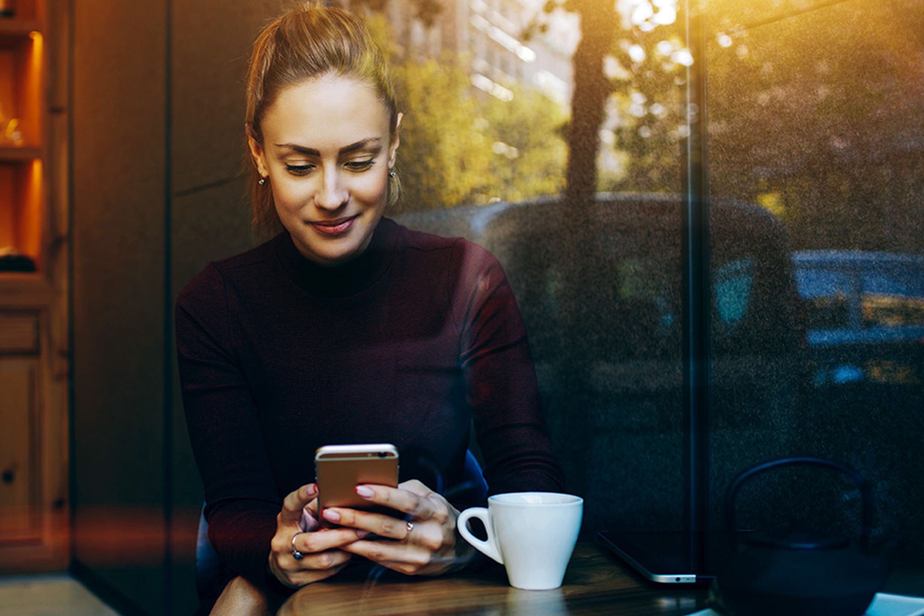 Woman sitting at a cafe table, looking at her phone, with a cup of coffee and window reflections of trees and buildings in the background.