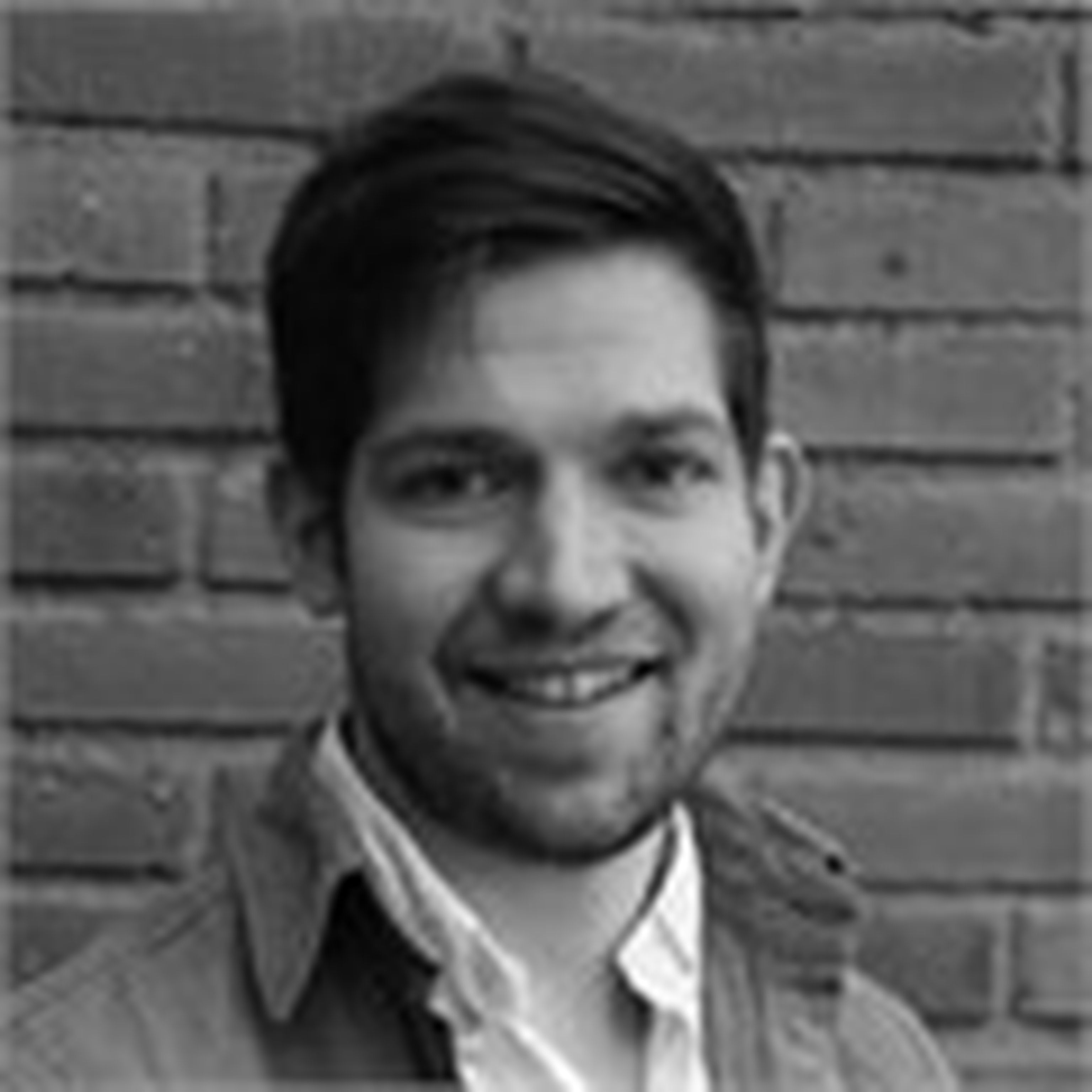 Black and white portrait of a smiling man with dark hair standing against a brick wall.