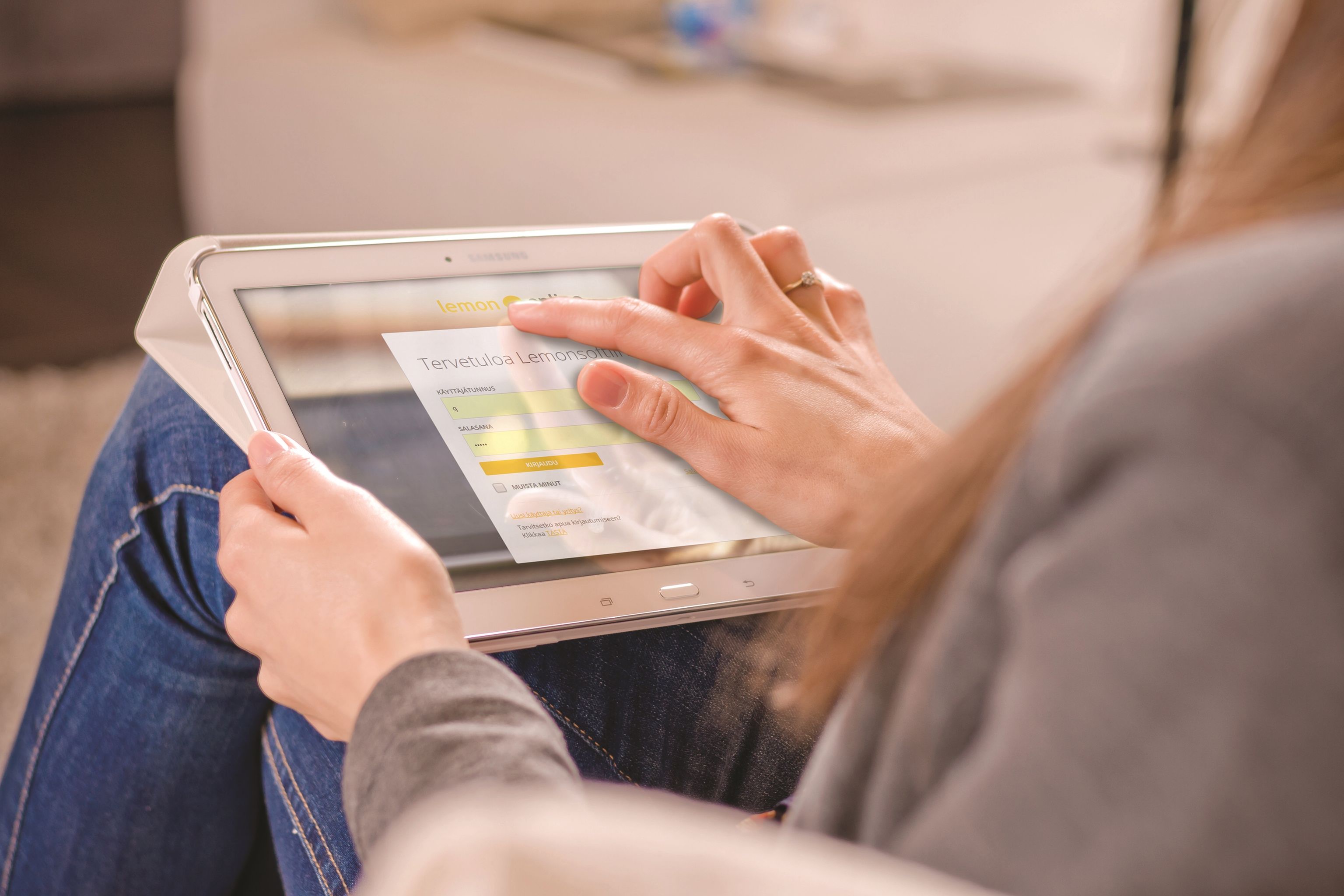 Henkilö käyttää tablettia ja tarkastelee sen näytöllä olevaa taloudellista tai tilastollista graafia, istuen sohvalla harmaa neule ja farkut yllään.