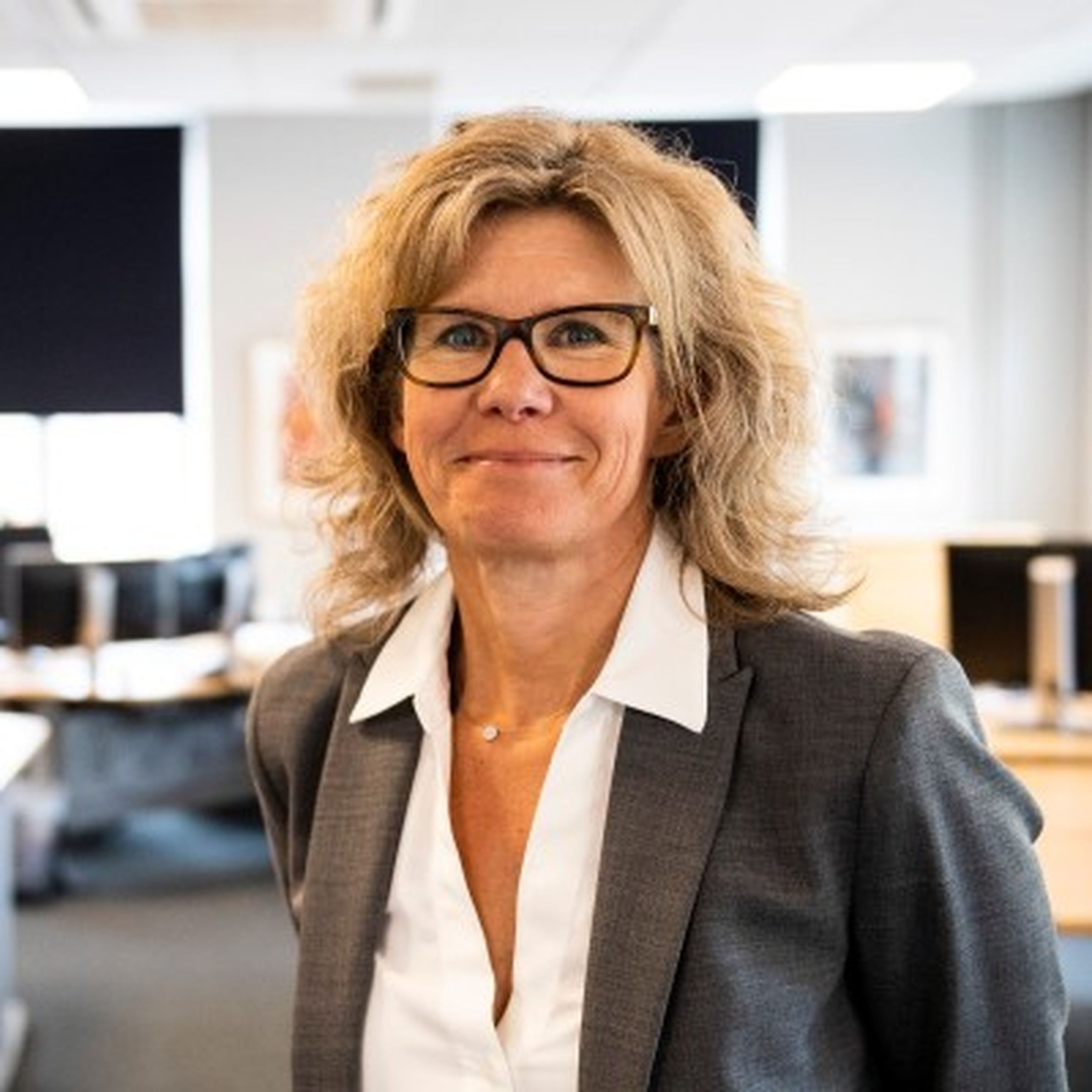 Profesjonell kvinne med blondt, bølgete hår og briller, iført en grå blazer og hvit skjorte, smilende i et moderne kontormiljø.