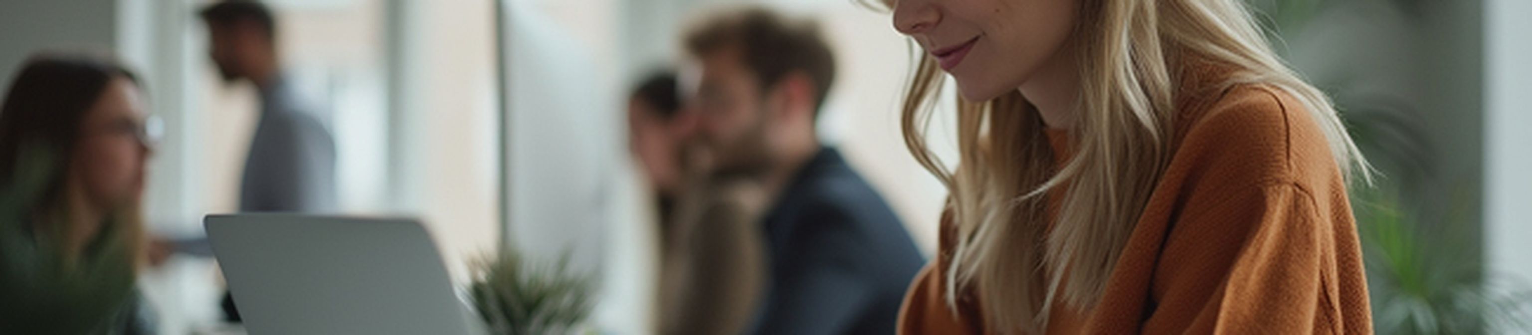 Woman in a rust-colored sweater working on a laptop at a bright, modern office with colleagues in the background.