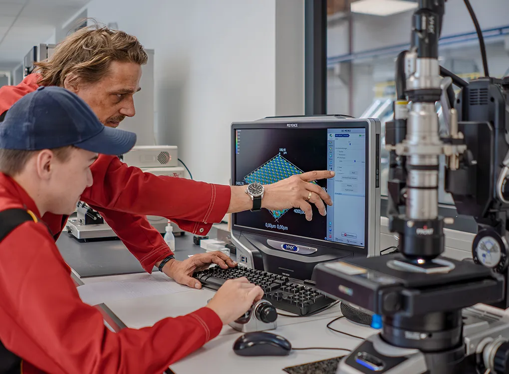 Two technicians in red uniforms analyze 3D data on a computer screen in a lab, with scientific equipment nearby.