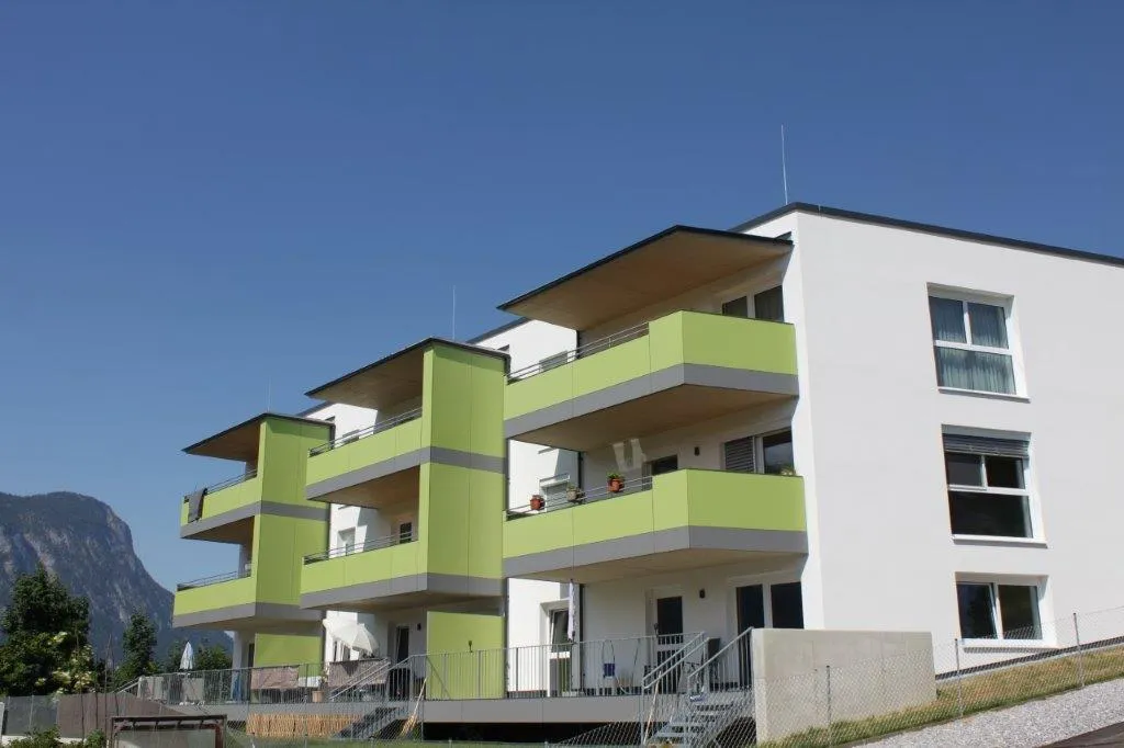 Modern white apartment building with green balconies set against a clear blue sky and mountain backdrop.