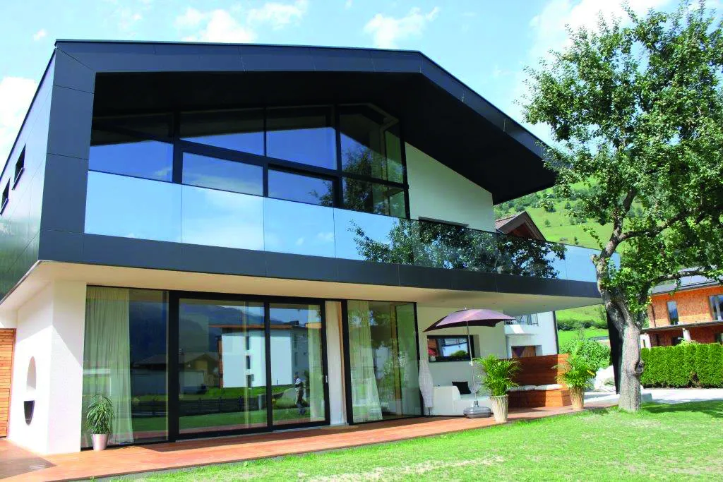 Modern two-story house with large glass windows, a balcony, and a spacious yard, under a blue sky with a few clouds.