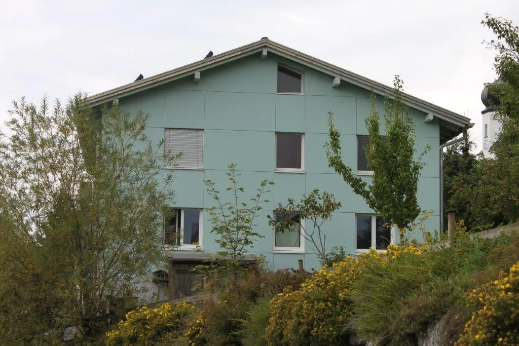 A light blue house with multiple windows, surrounded by trees and bushes, set on a grassy slope with a cloudy sky above.
