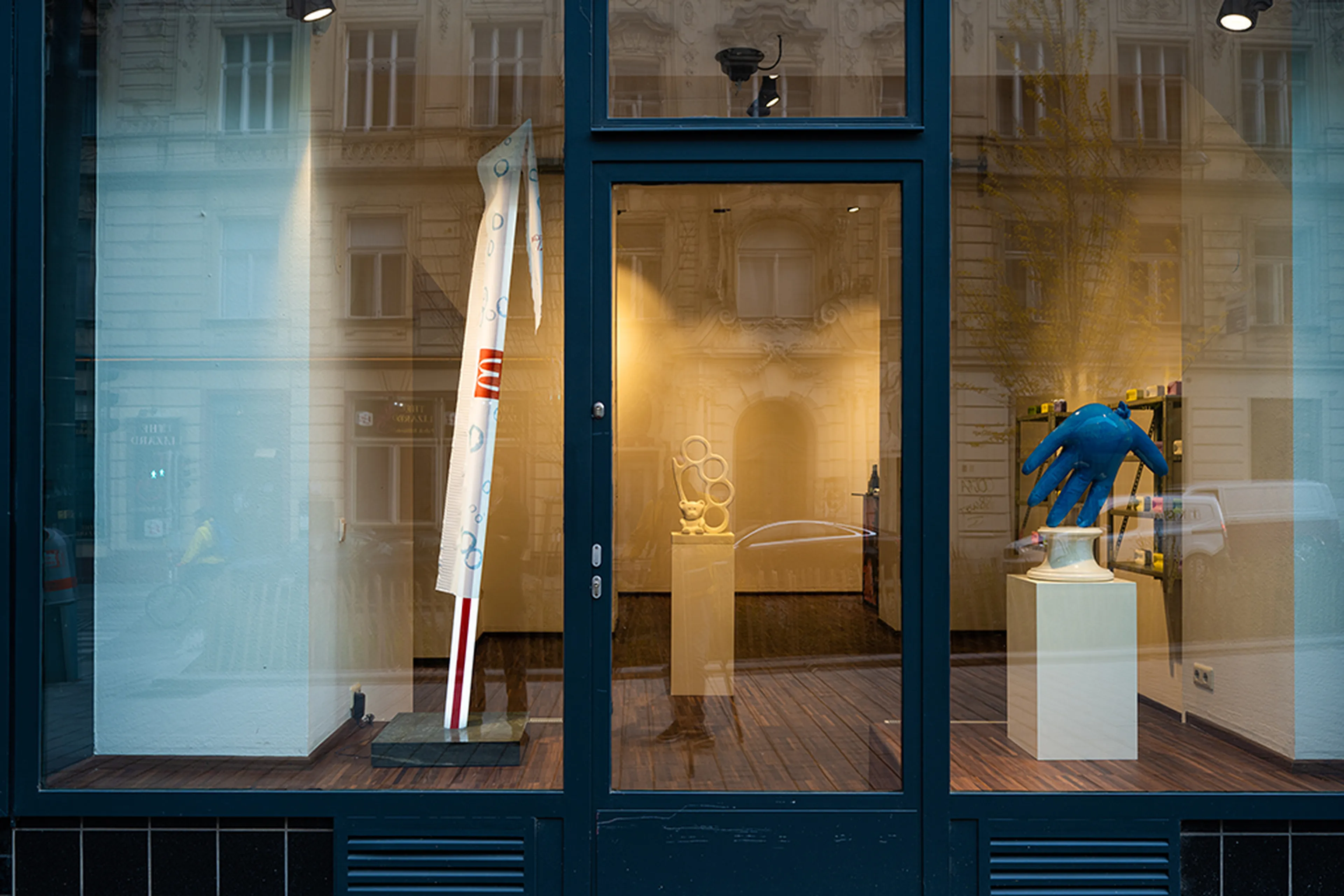 Art gallery window display featuring abstract sculptures, including a tall white piece and a large blue hand, against a wooden floor backdrop.