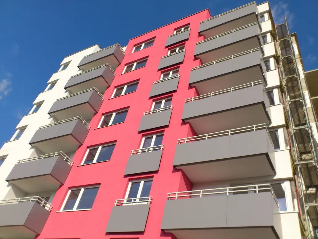 A modern apartment building with a vibrant pink and white facade, featuring multiple balconies and set against a clear blue sky.