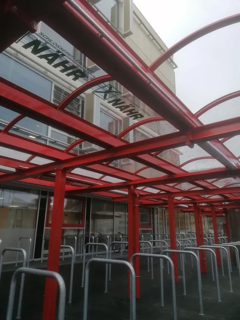 Red metal bike racks with a transparent arched roof outside a building, featuring glass windows and signage overhead.