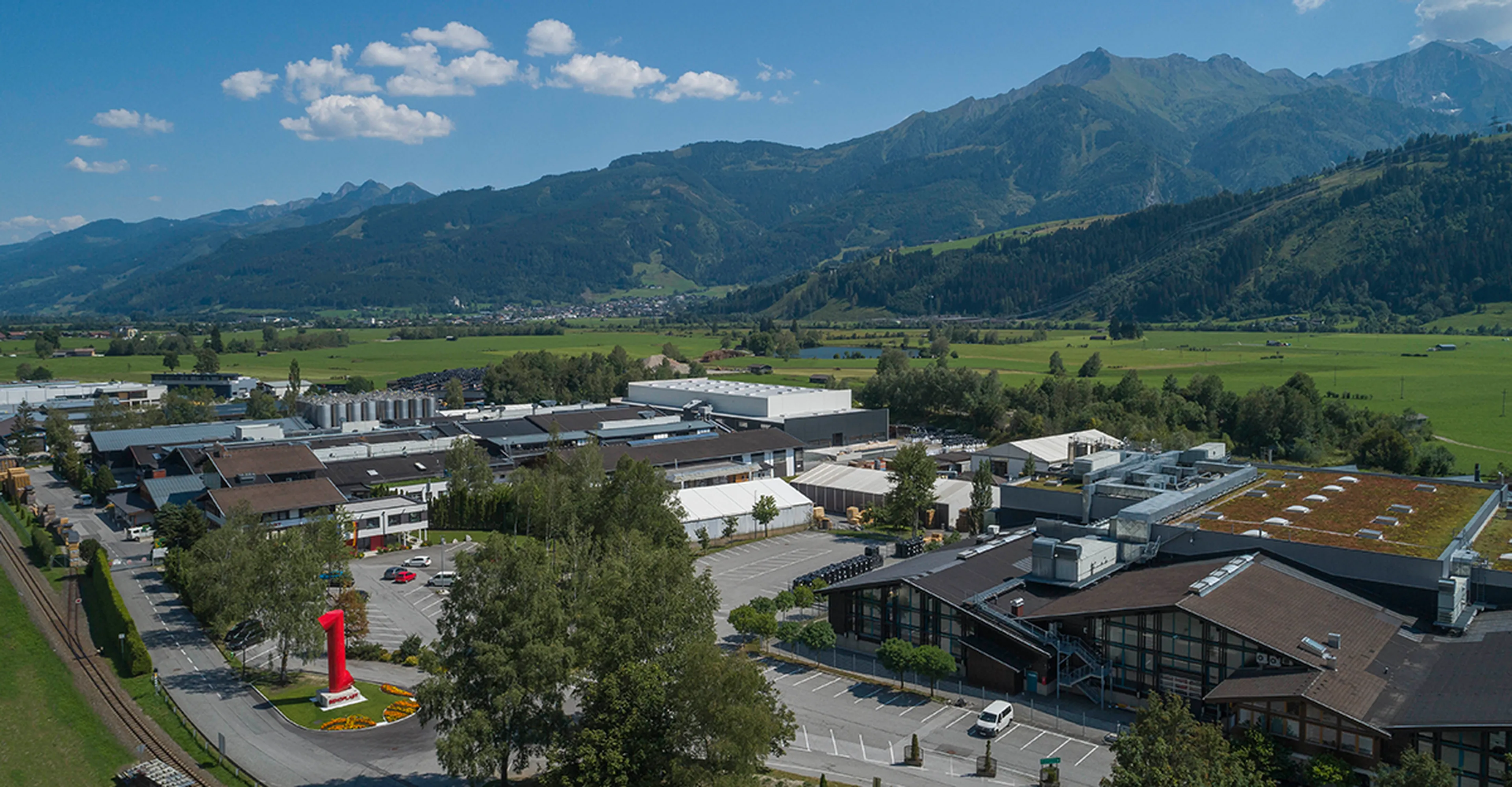 Aerial view of an industrial area with a large red number one sculpture, surrounded by mountains and green fields under a blue sky.