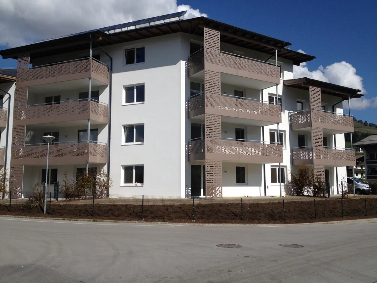Modern four-story apartment building with white walls, brown balconies, and a sloped roof, under a blue sky with clouds.