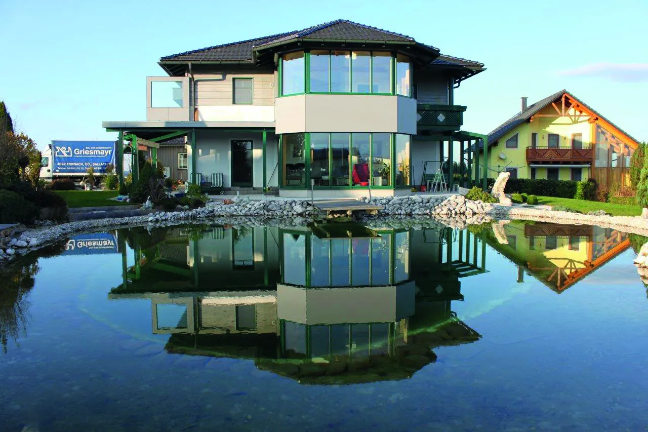 Modern two-story house with glass facade reflecting in a tranquil pond, surrounded by greenery and a clear blue sky.