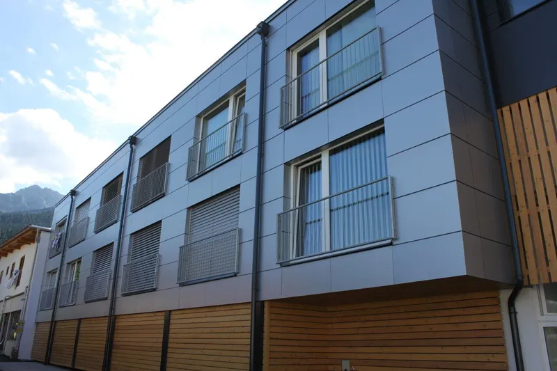 Modern building with metal paneled facade, multiple windows with shutters, and wooden lower section, set against a partly cloudy sky.