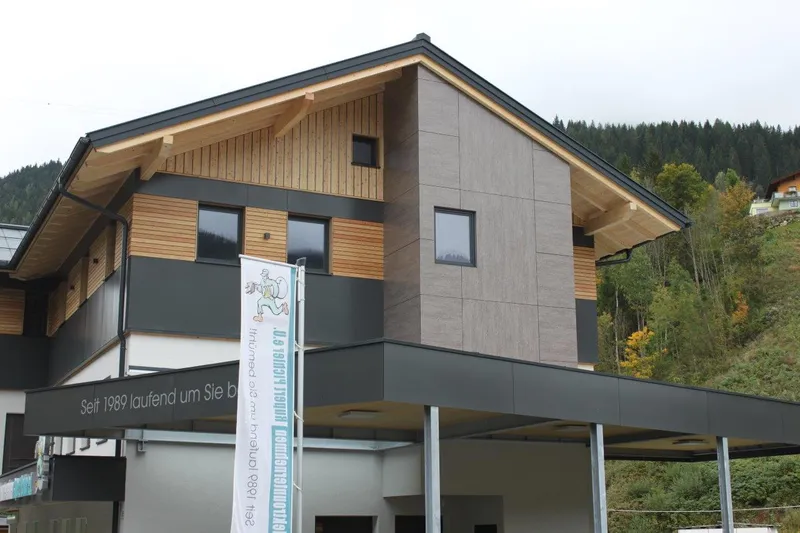 Modern two-story building with a wooden facade and large windows, set against a backdrop of trees and hills under a cloudy sky.