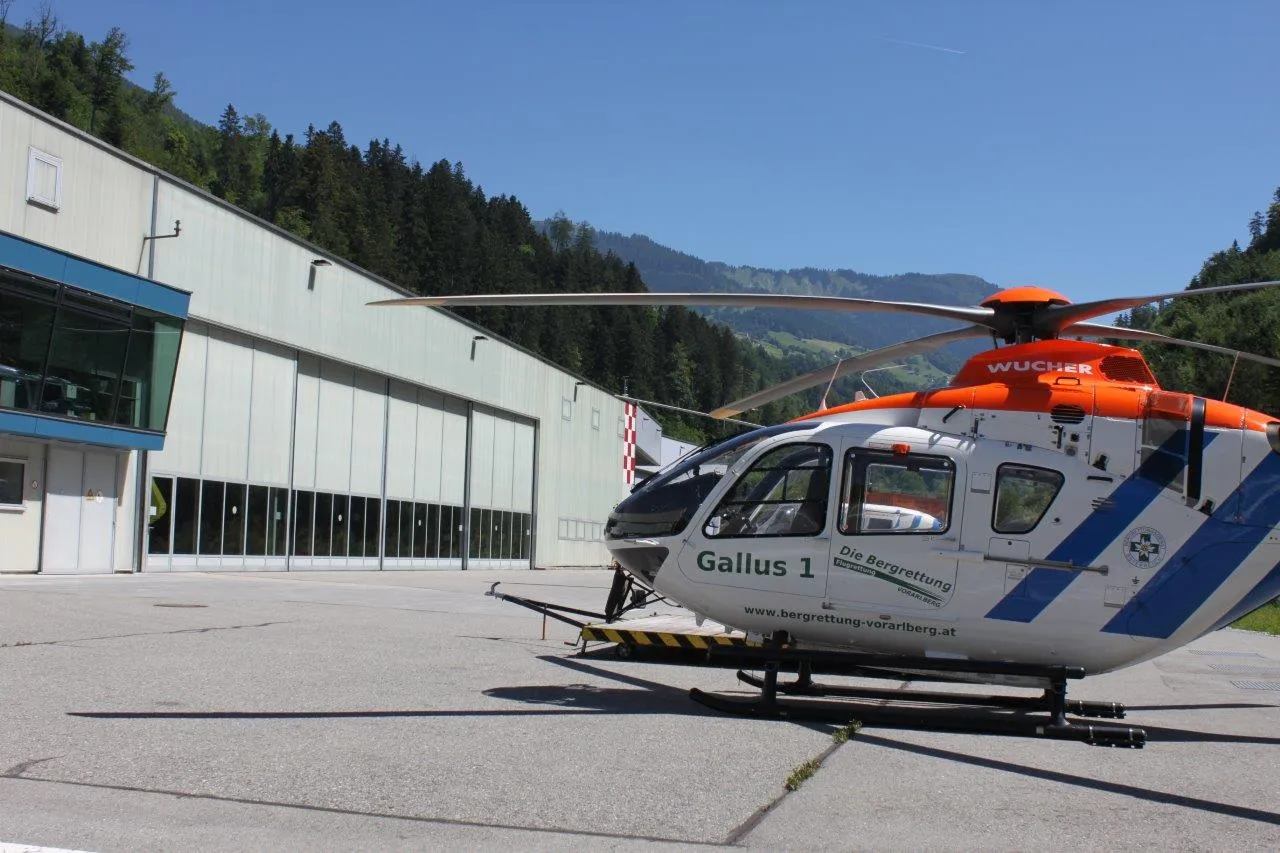 A rescue helicopter is parked on a concrete surface near a large building, with forested hills and mountains in the background.
