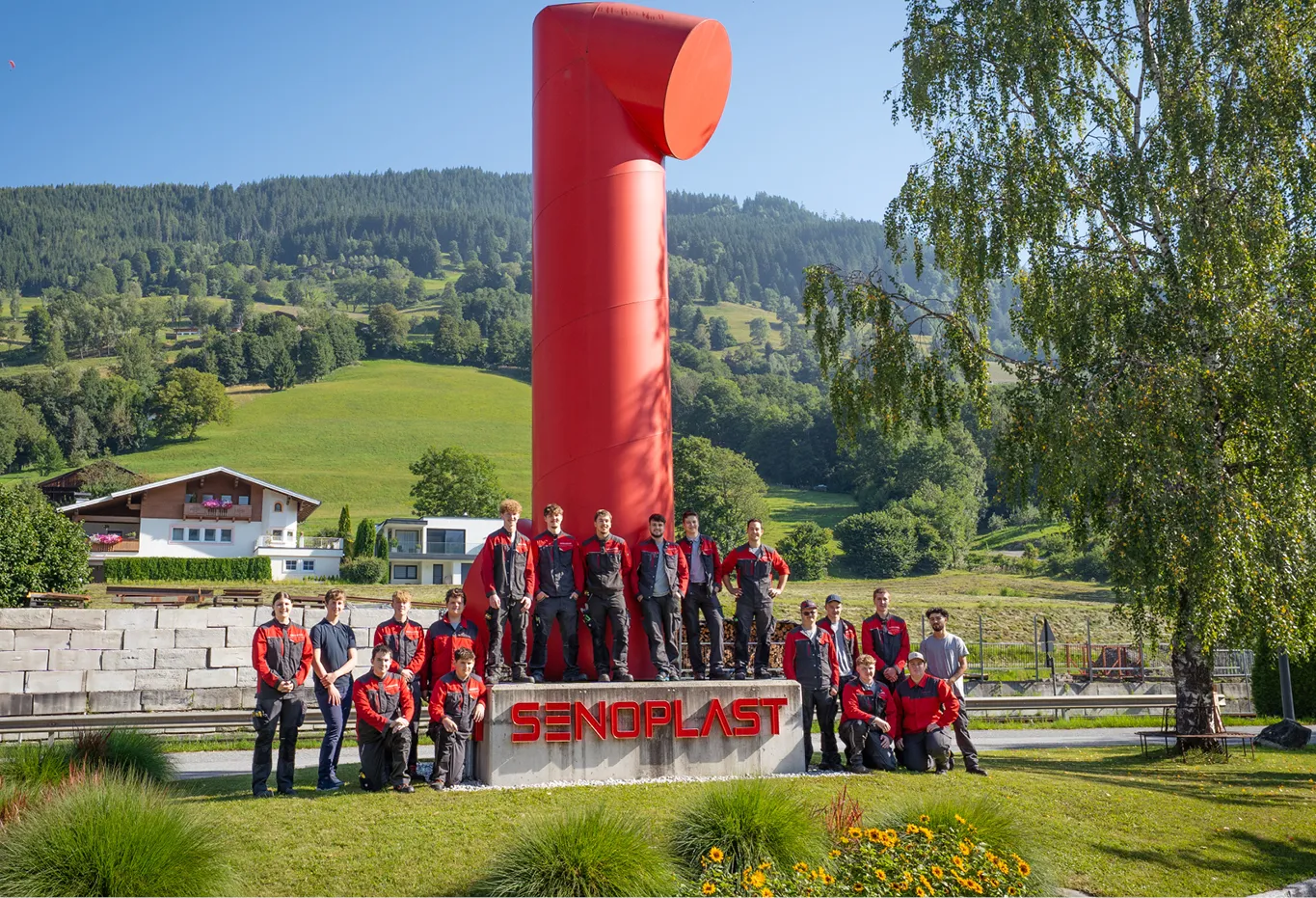 Group of people in matching red and black outfits pose around a large red sculpture with "Senoplast" sign, set against a scenic mountain backdrop.