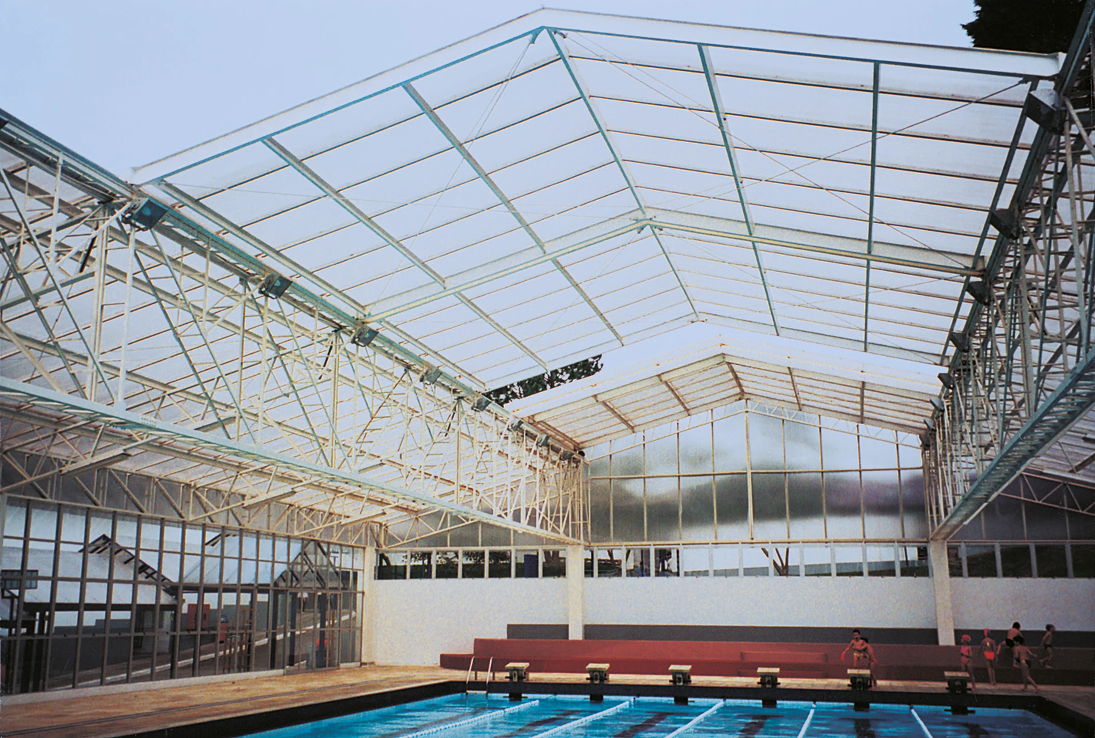 Indoor swimming pool with a high, transparent ceiling and large windows. A few people are seated on benches by the poolside.