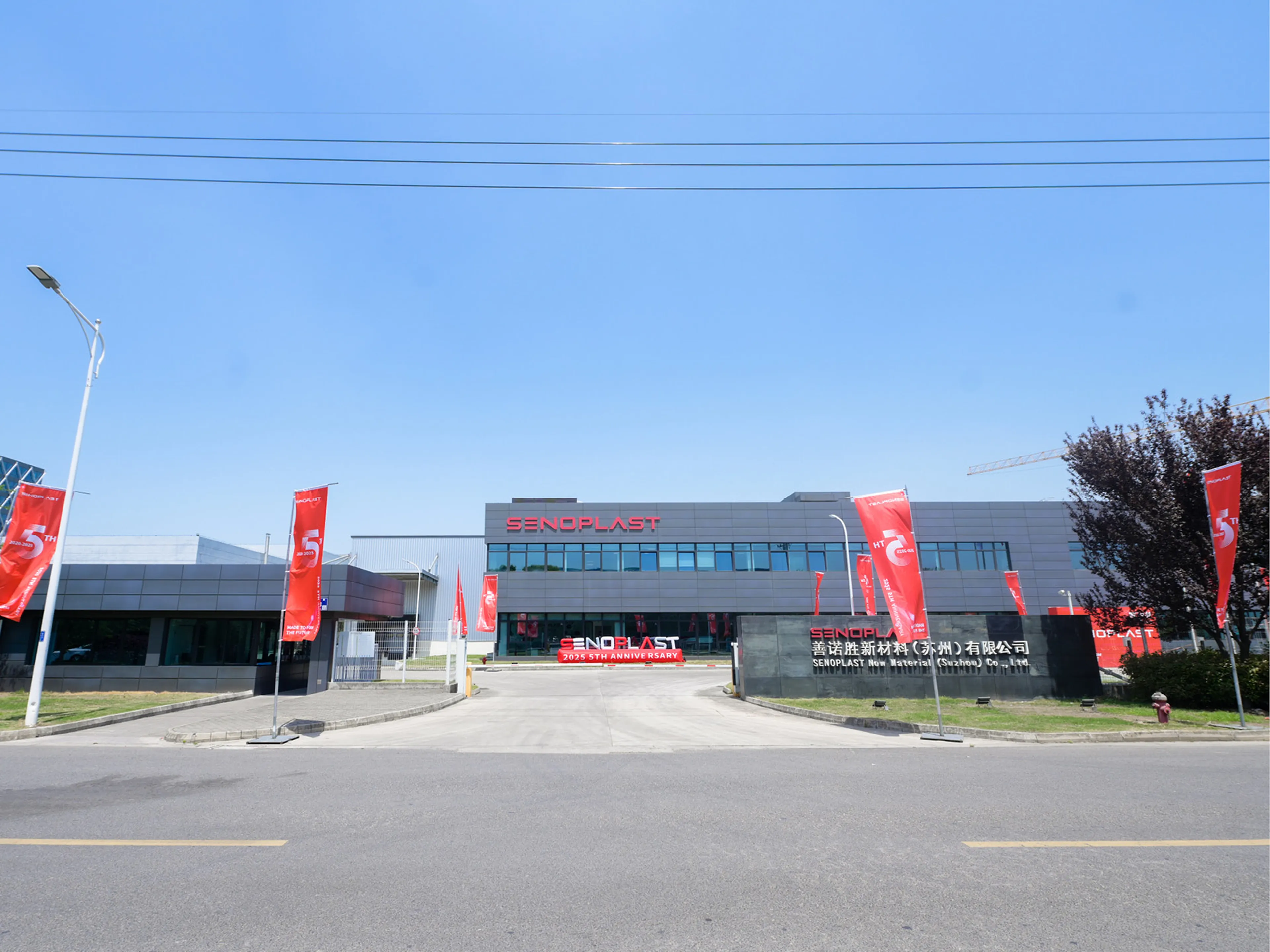 Modern industrial building with "SENOPLAST" signage, red flags lining the driveway, and clear blue sky in the background.