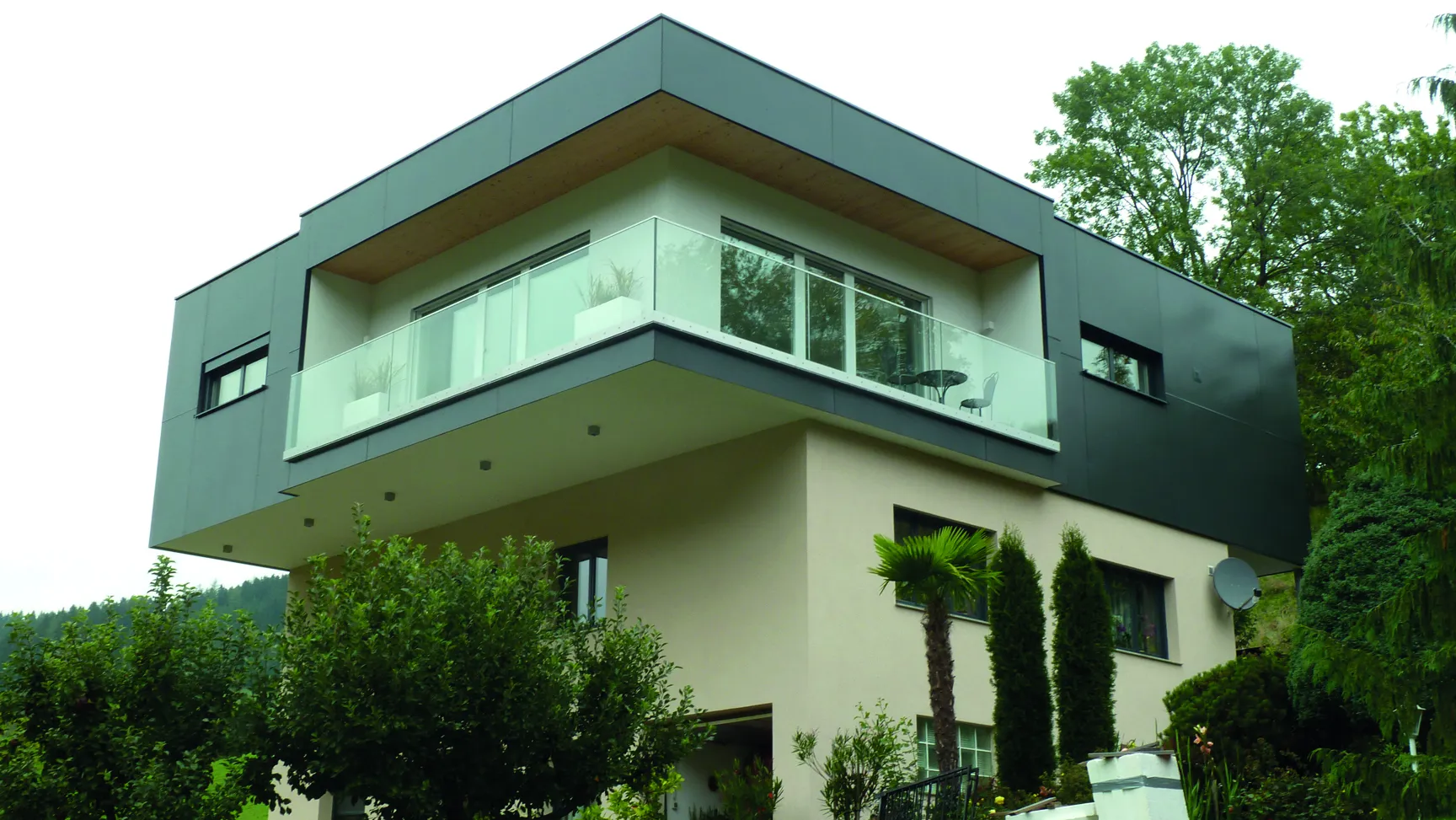 Modern two-story house with a dark upper facade, large windows, and a balcony, surrounded by trees and greenery.