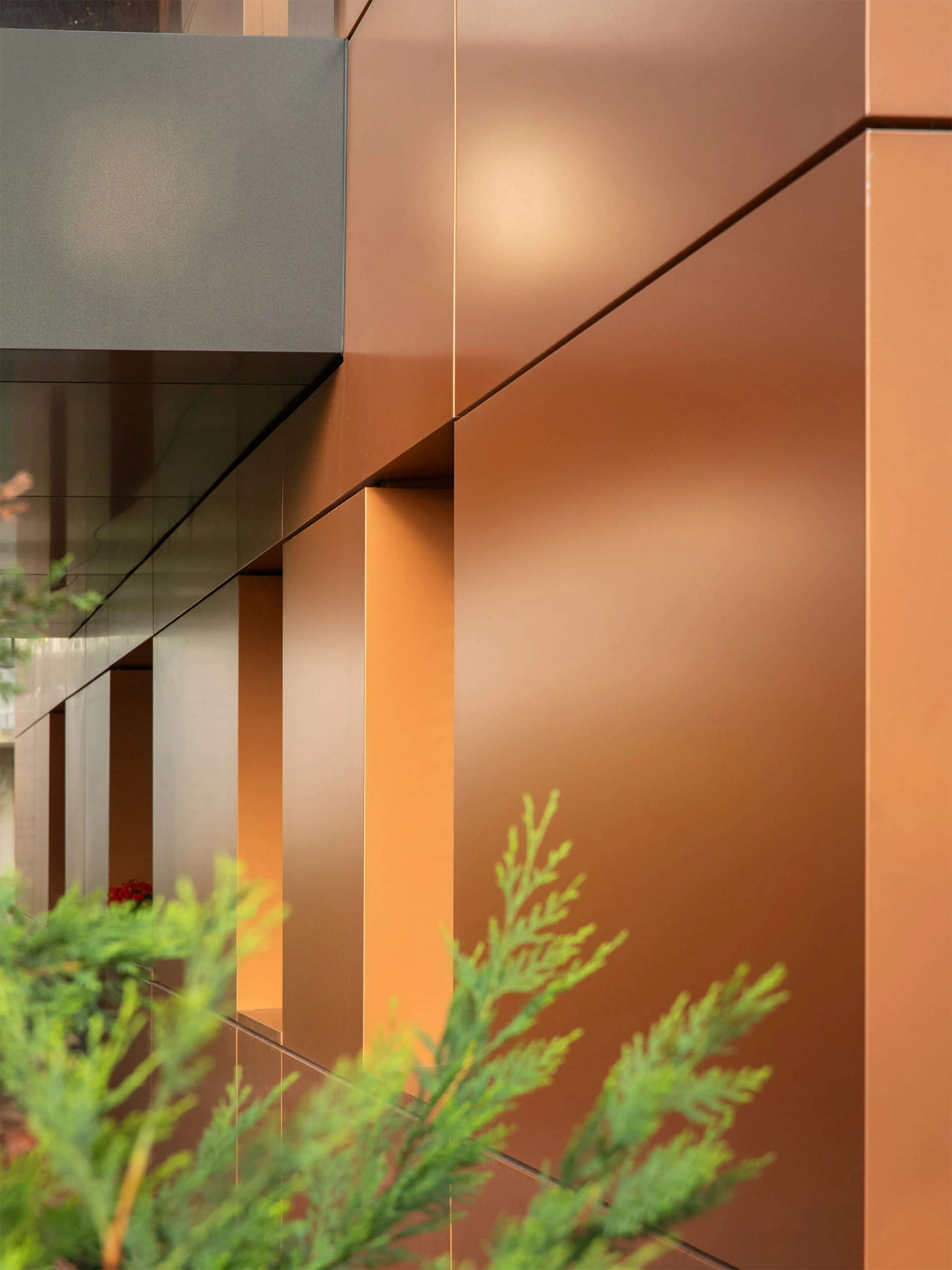 Modern building facade with smooth, copper-colored panels and a contrasting dark section, with greenery in the foreground.