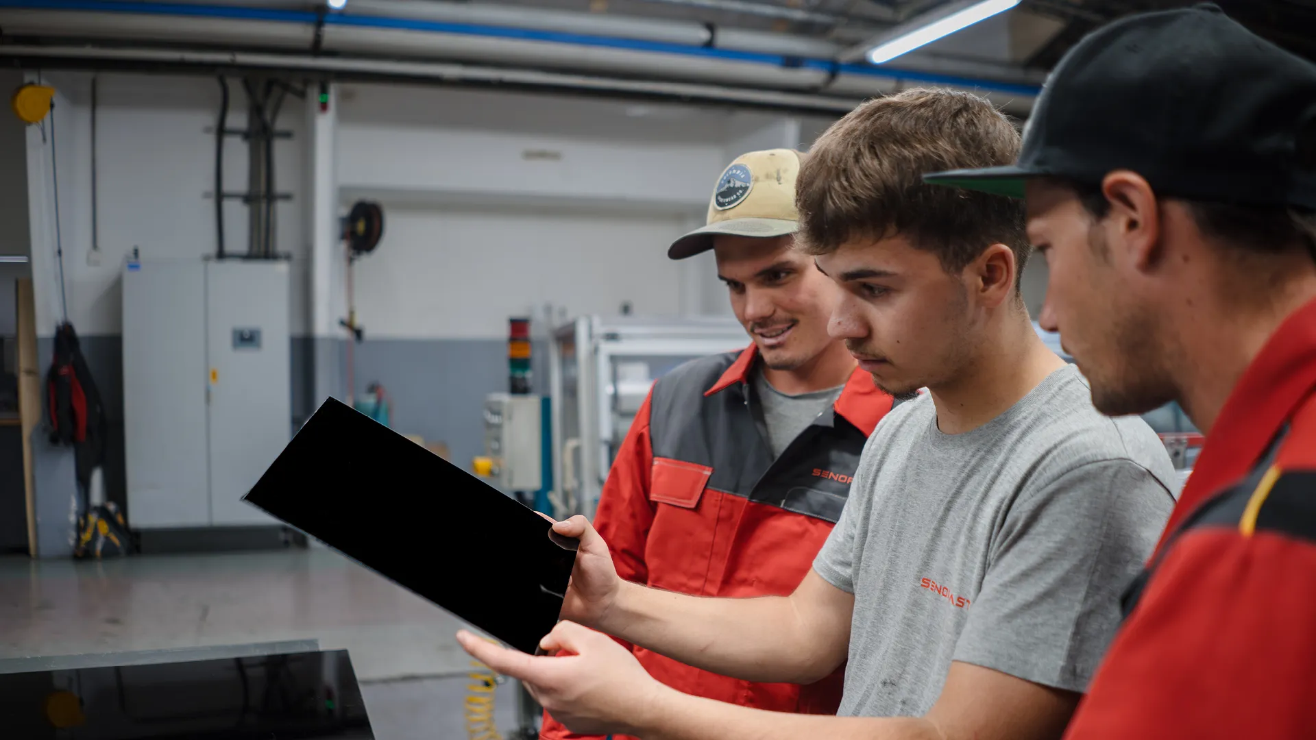 Three men in a workshop examining a black panel. One holds it while the others observe, surrounded by industrial equipment.