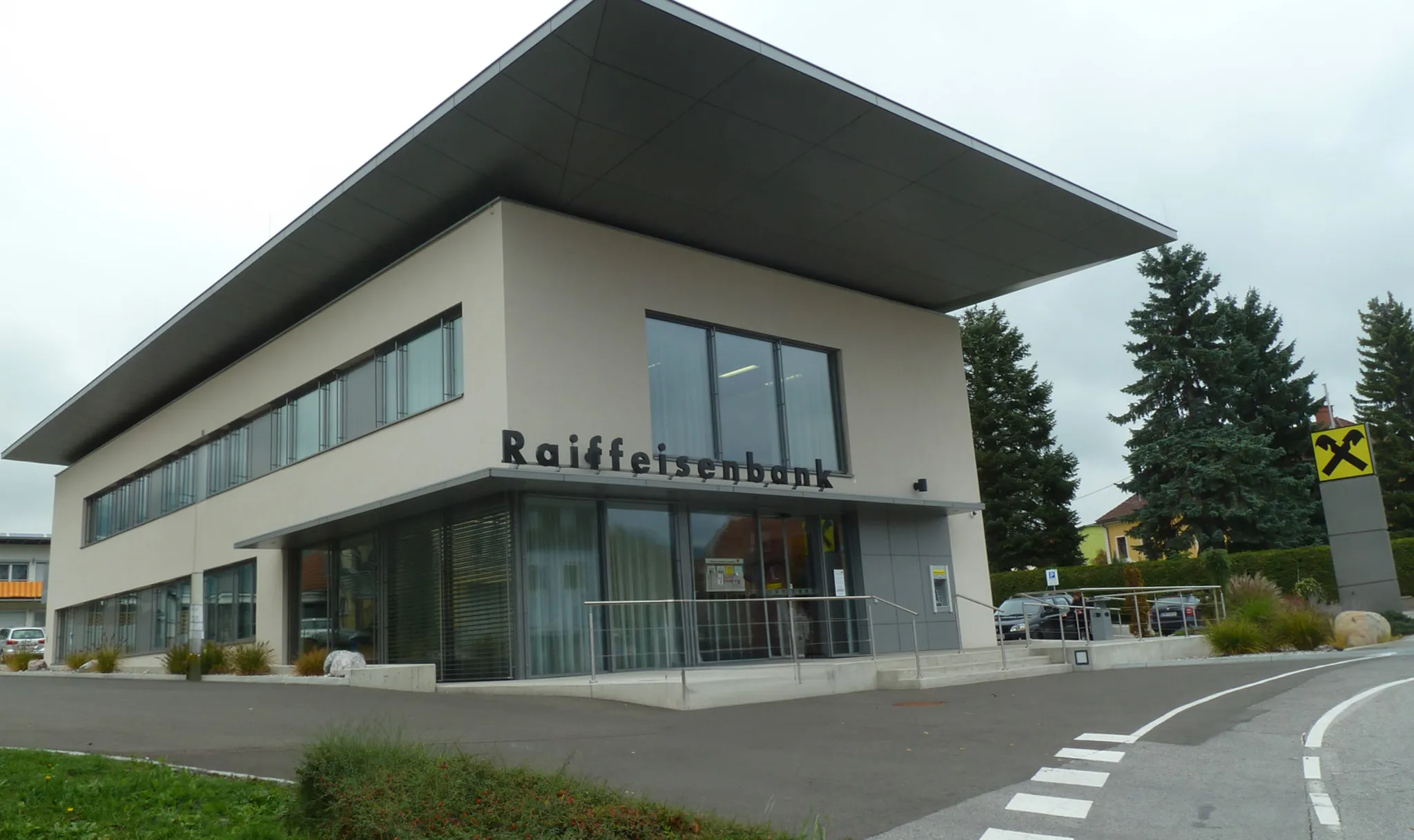 Modern two-story bank building with large windows, labeled "Raiffeisenbank," situated on a corner lot with a pathway and trees nearby.