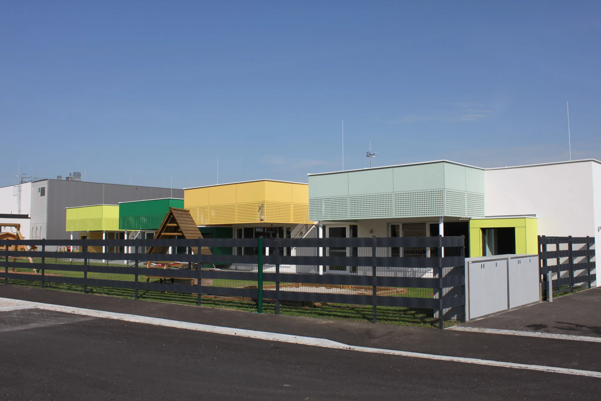 Modern building with colorful sections, including green, yellow, and white, surrounded by a black fence under a clear blue sky.
