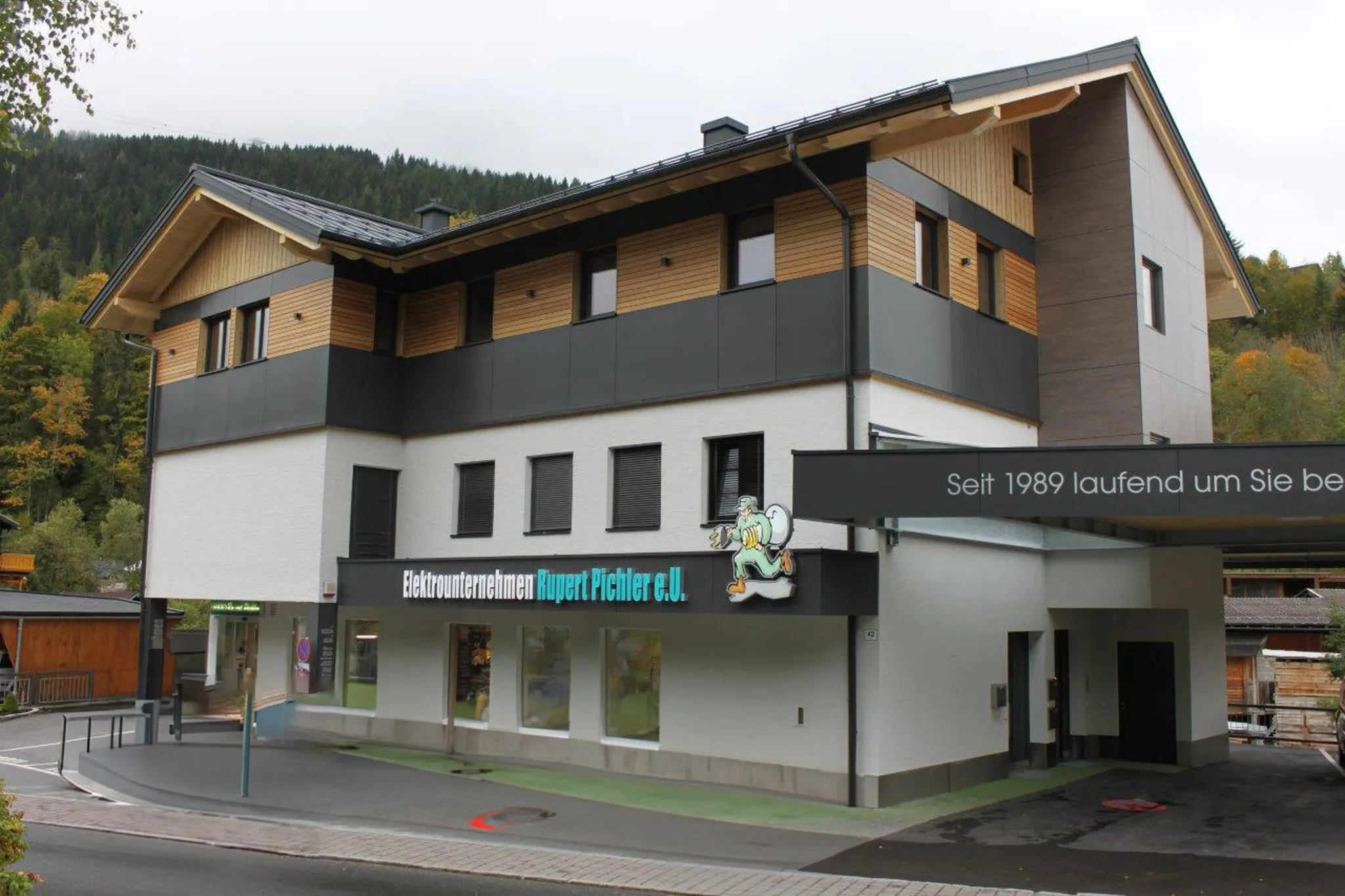 Modern multi-story building with signage, surrounded by trees and mountains, featuring large windows and a sloped roof.