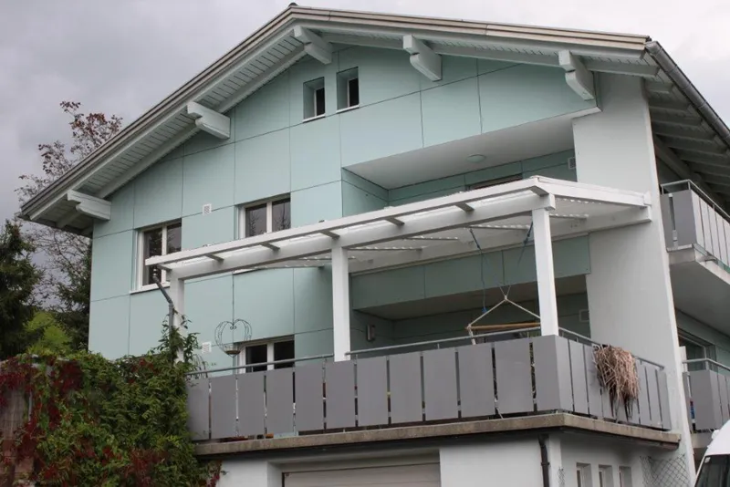 Light blue house with a balcony covered by a white pergola. Grey railings and overcast sky in the background. Ivy grows on the wall.