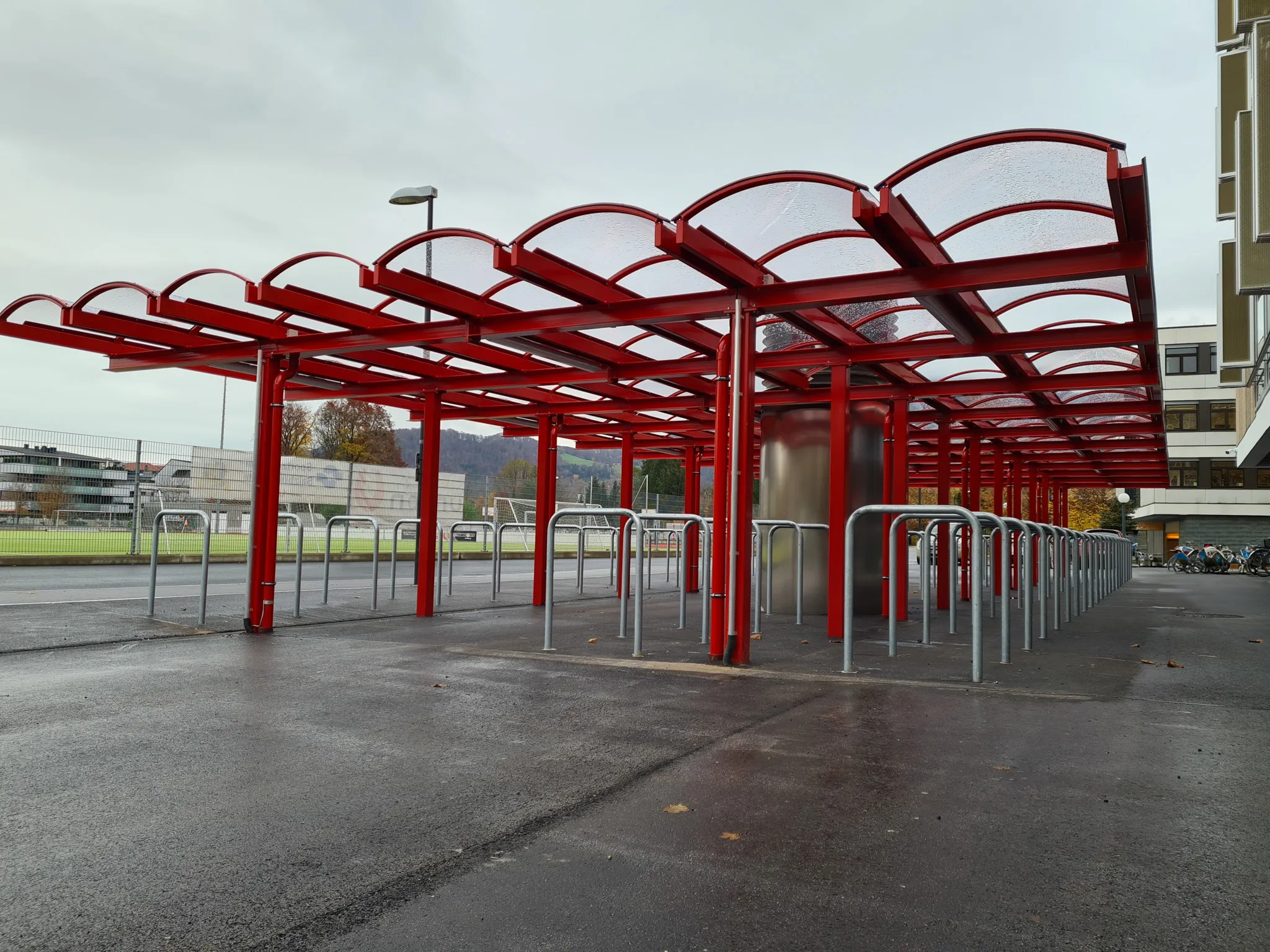 Red metal bike shelter with a curved roof and empty racks, situated on wet pavement near a building and sports field under a cloudy sky.
