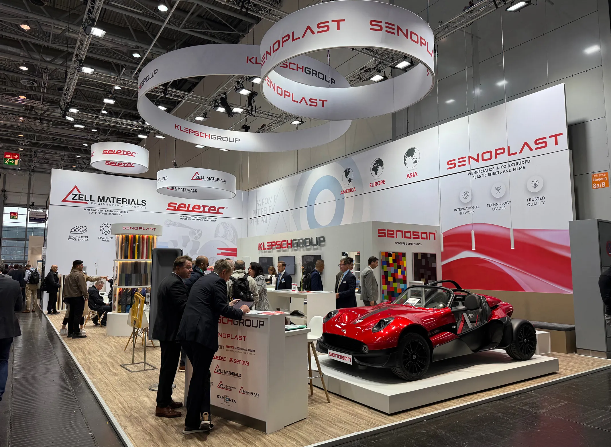 Trade show booth with Senoplast signage, hanging circular banners, visitors at counters and a glossy red open-top concept car on display.