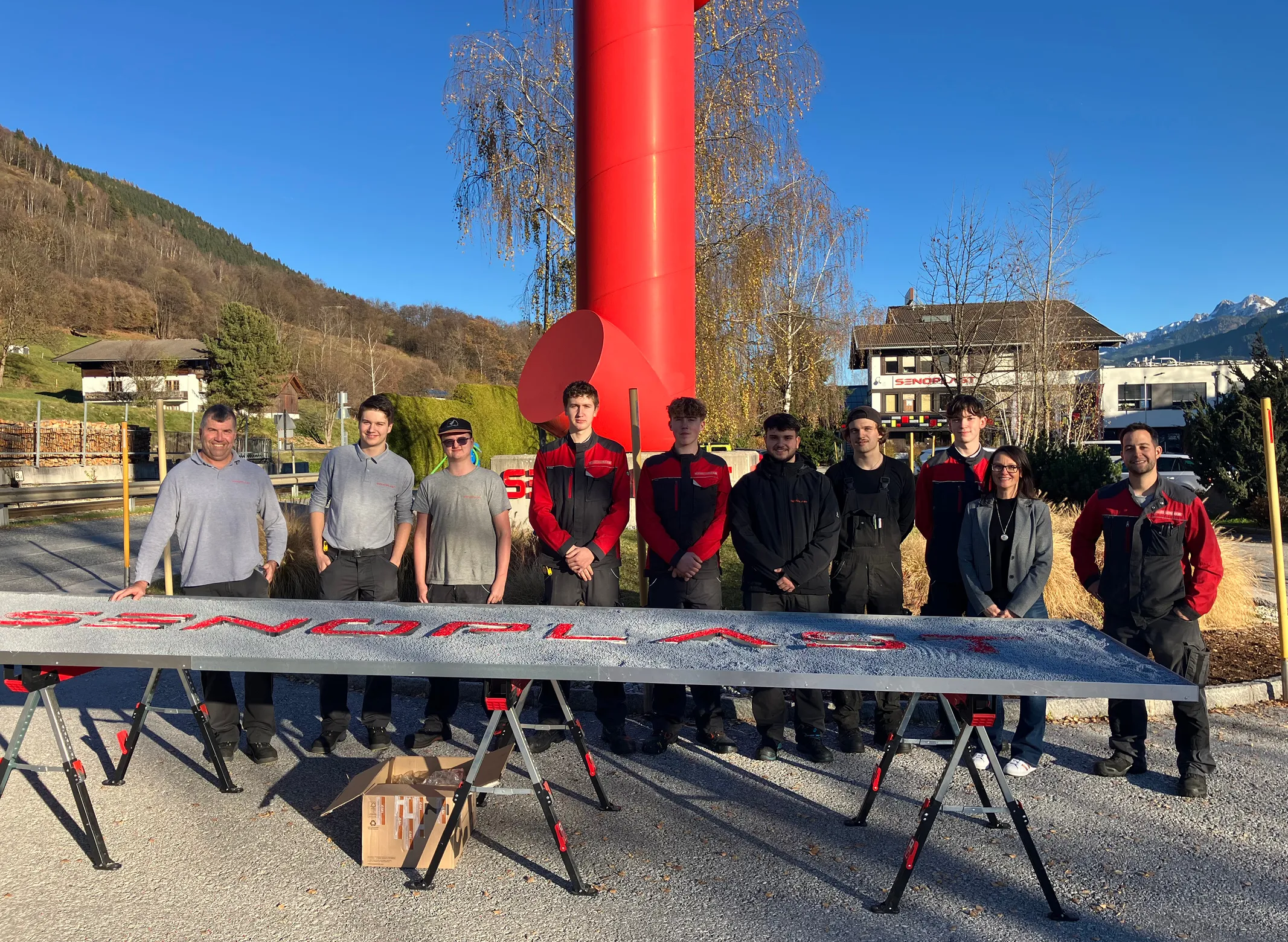 A group of nine people standing outdoors in front of a large red structure, with mountains and buildings in the background.