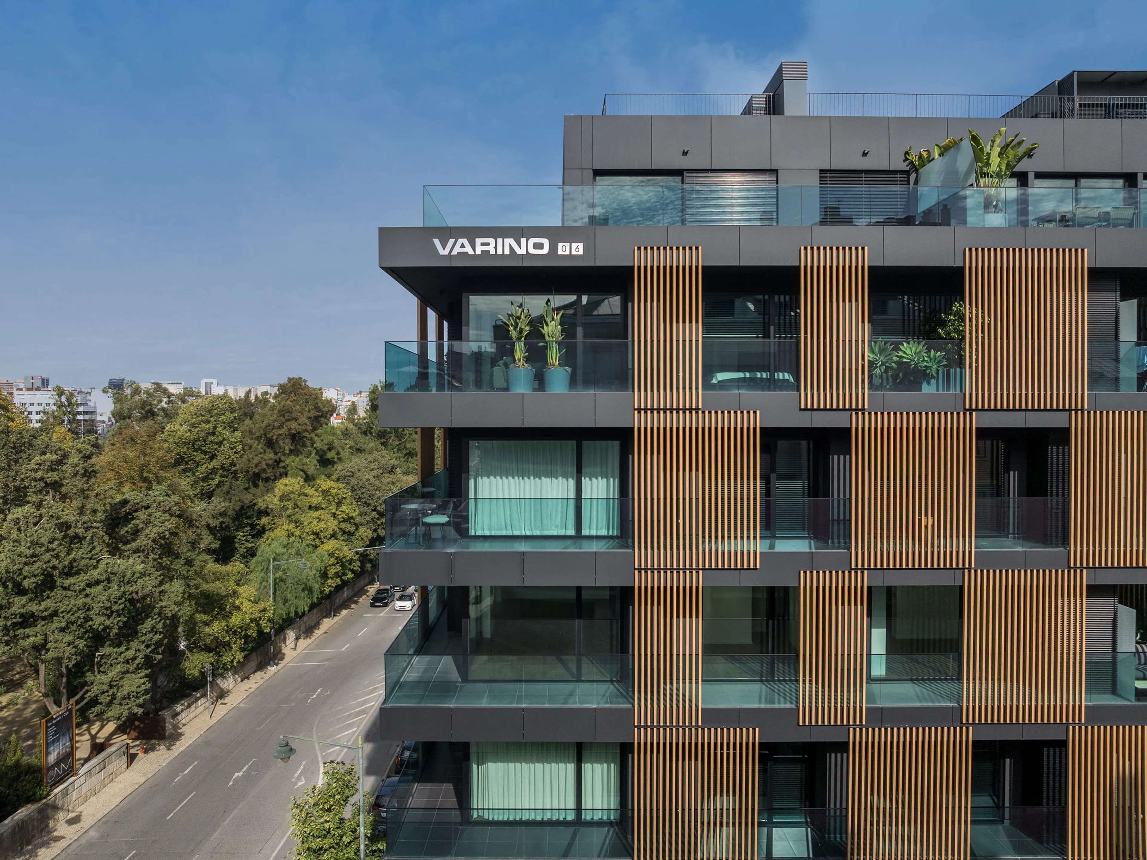 Modern building with wooden slat facade, glass balconies, and greenery. Situated beside a tree-lined road under a clear blue sky.