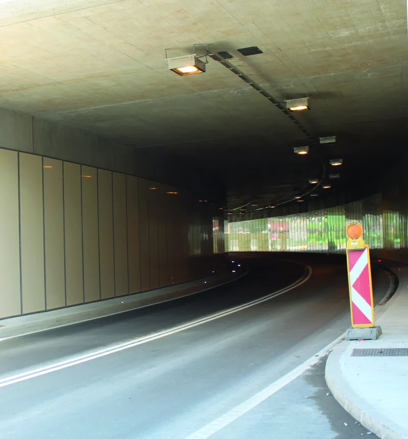 A dimly lit road tunnel with a curve, featuring a warning sign on the right and a paneled wall on the left.