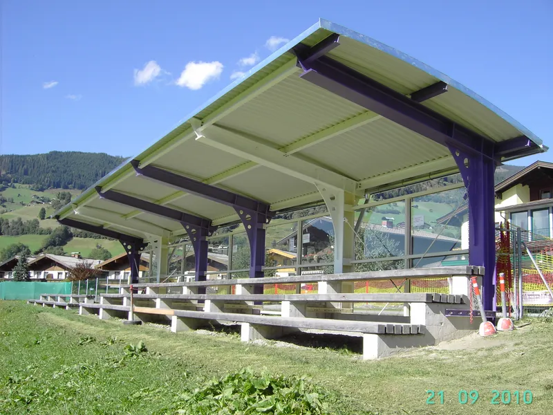 Von SENO Spezialmaschinen gefertigte Tribüne am Fußballplatz in Piesendorf mit Metalldach und Holzsitzen, eingebettet in eine Rasenfläche mit Bergen und Häusern im Hintergrund.