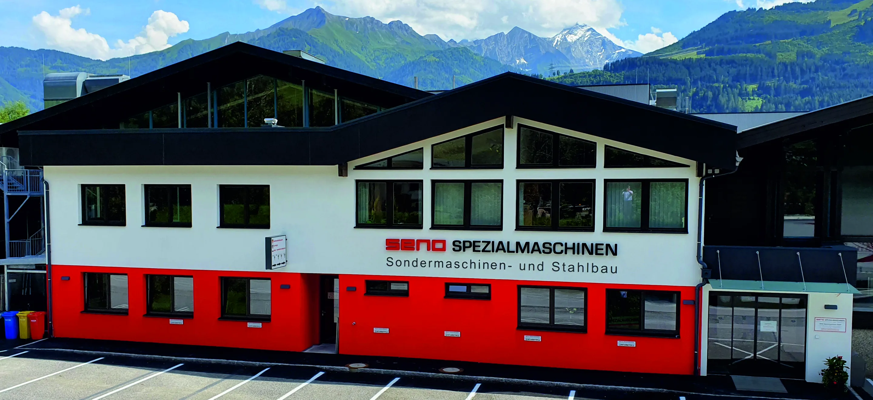 Frontansicht des Bürogebäudes von SENO Spezialmaschinen, das Erdgeschoss mit seiner roten Fassadenfarbe hebt sich vom grün-grau-weißen Hintergrund der bewaldeten Hügel und schneebedeckten Berge ab