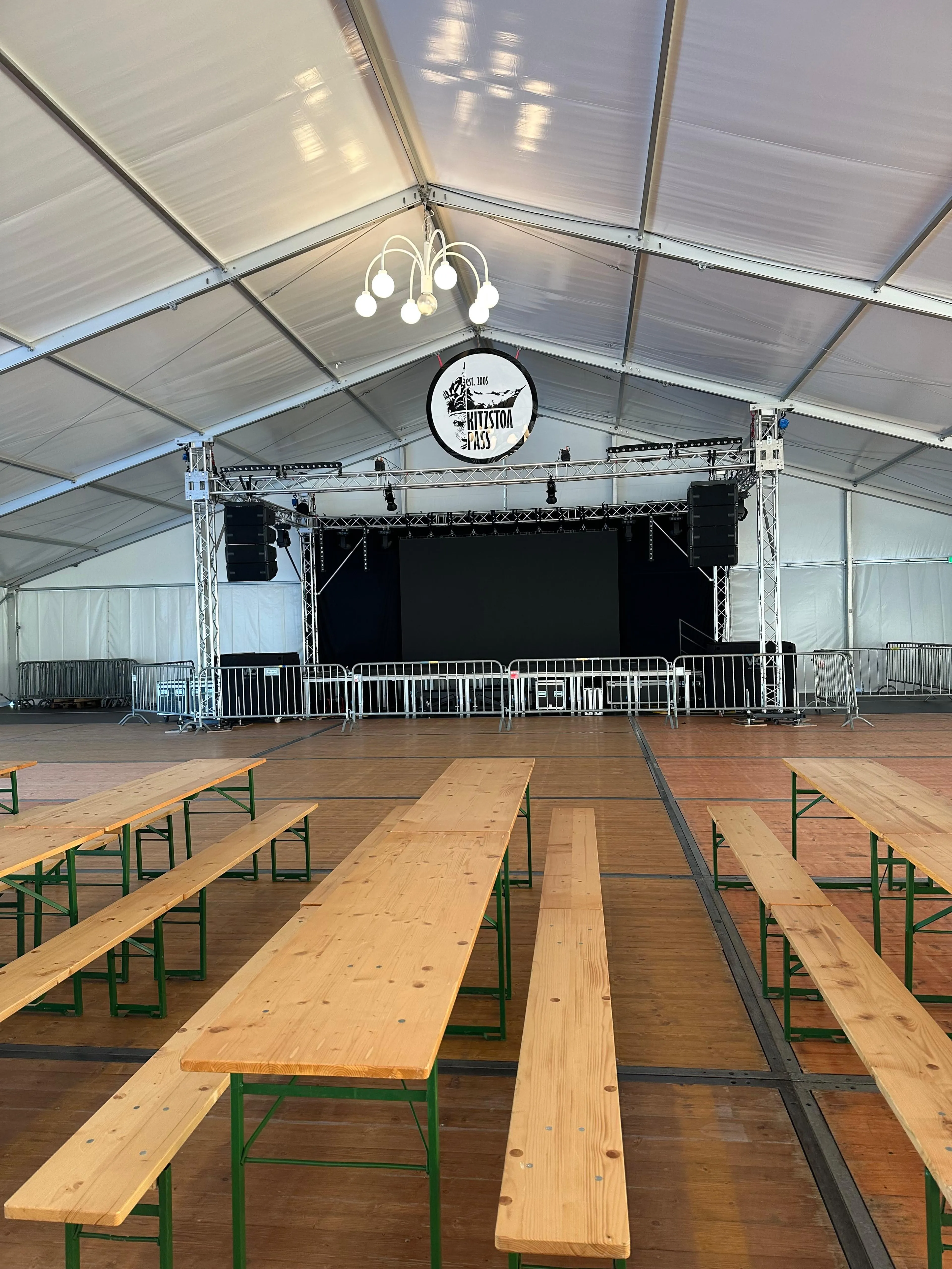 Großes Veranstaltungszelt mit Bühne, Holzbänken und Tischen. Über der Bühne sind eine Deckenbeleuchtung und das runde Logo der Kitzstoa Pass zu sehen.
