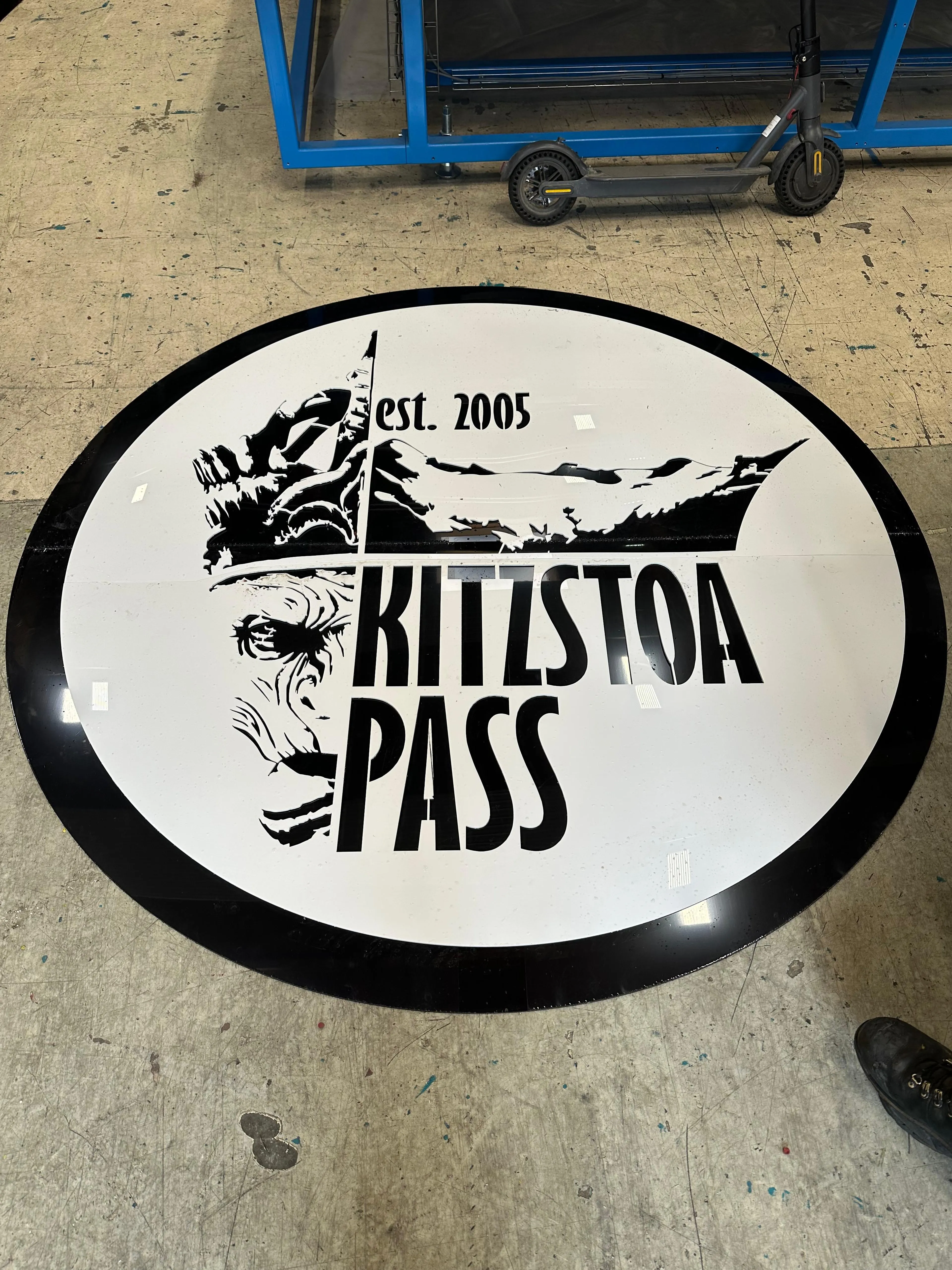 Rundes Logo auf dem Boden mit der Aufschrift „Kitzstoa Pass“ und „est. 2005“, mit einem stilisierten Berg- und Gesichtsdesign.