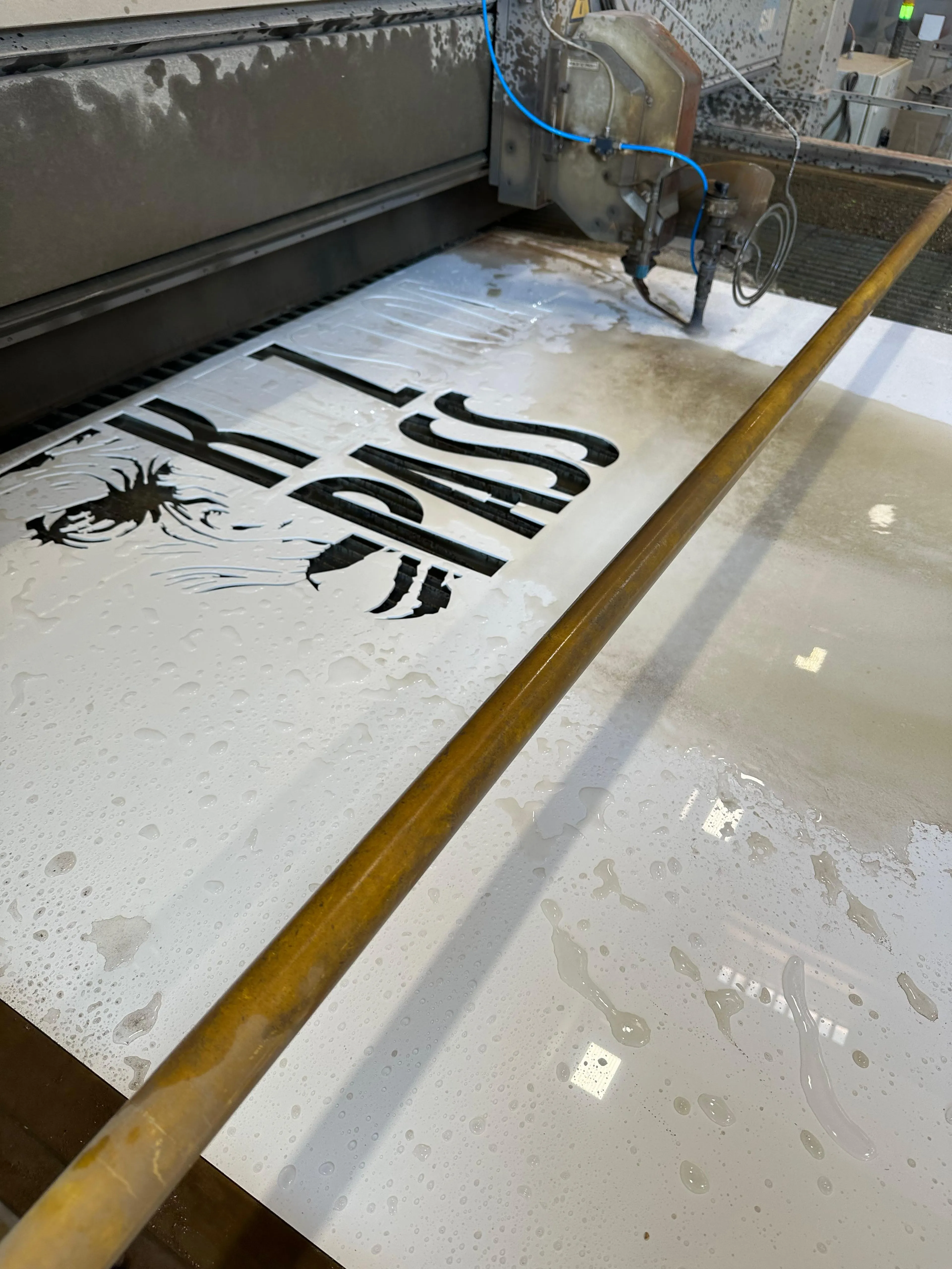 Eine Wasserstrahlschneidemaschine schneidet ein Design mit den Buchstaben „Kitzstoa PASS” mit stilisiertem Berg in ein Metallblech.