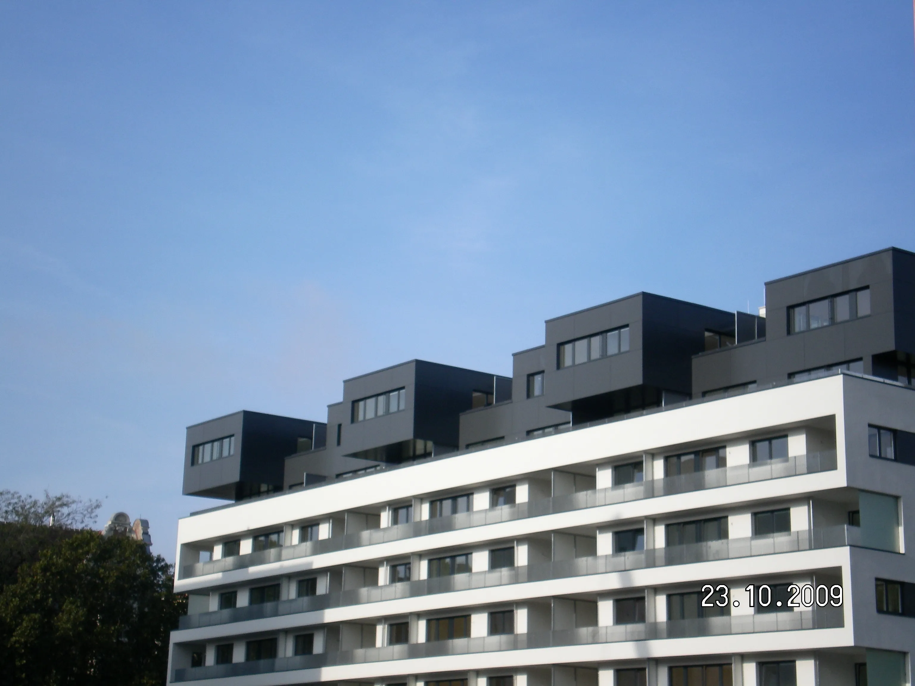 Modern multi-story building with staggered black and white balconies against a clear blue sky. Date stamp on the image reads 23.10.2009.