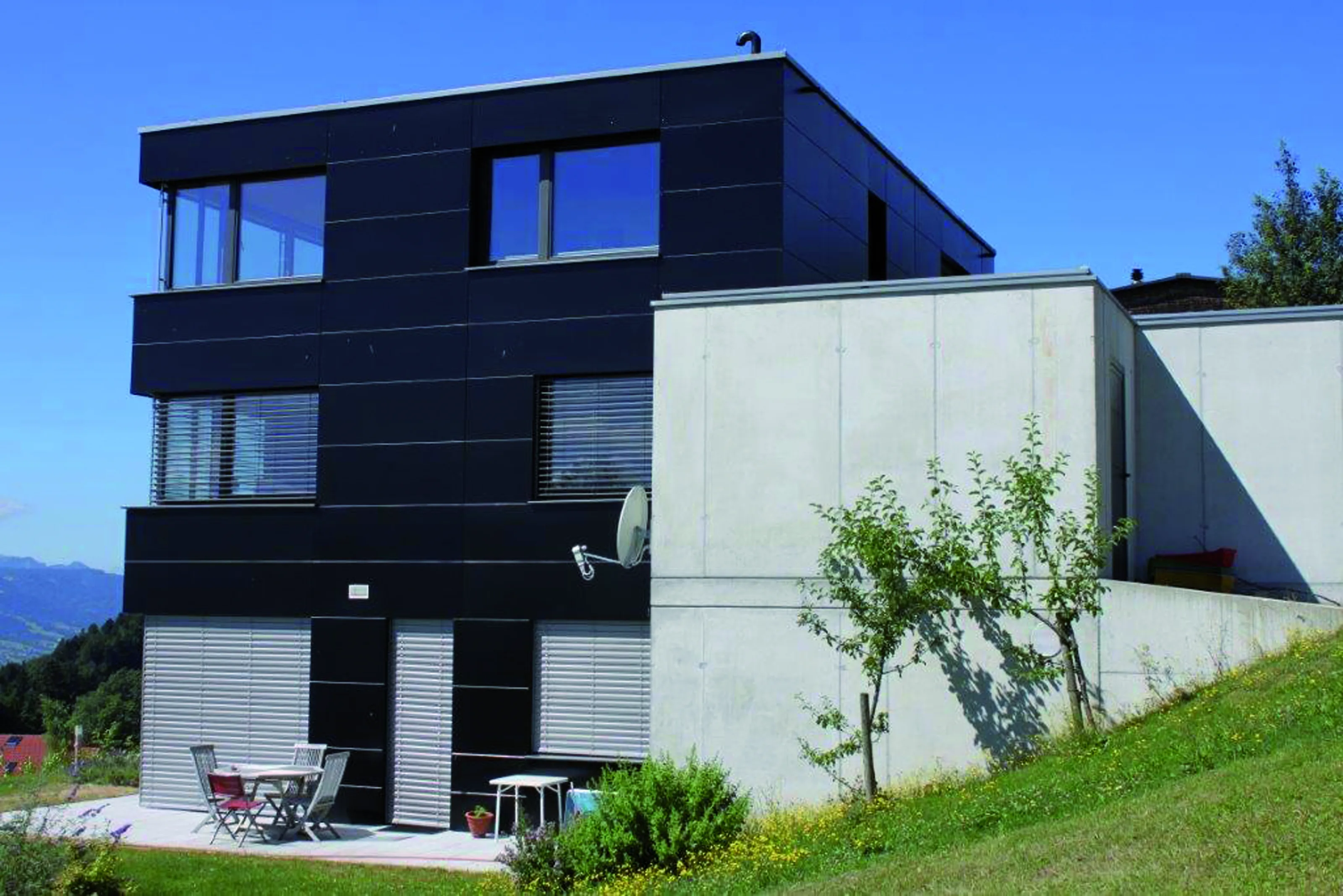 Modern three-story house with a black facade and large windows, adjacent to a concrete wall. Small patio with table and chairs, surrounded by greenery.
